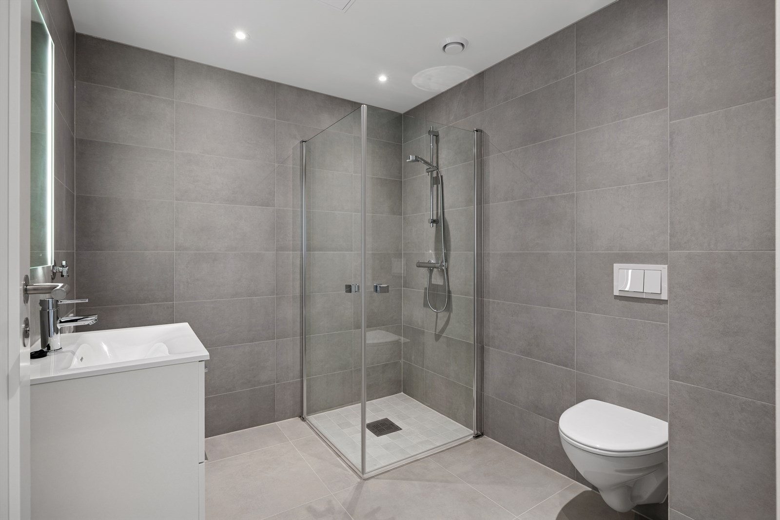 Moderne og flislagt bad med dusjhjørne, vegghengt toalett og servant med underskap. Galleribilde