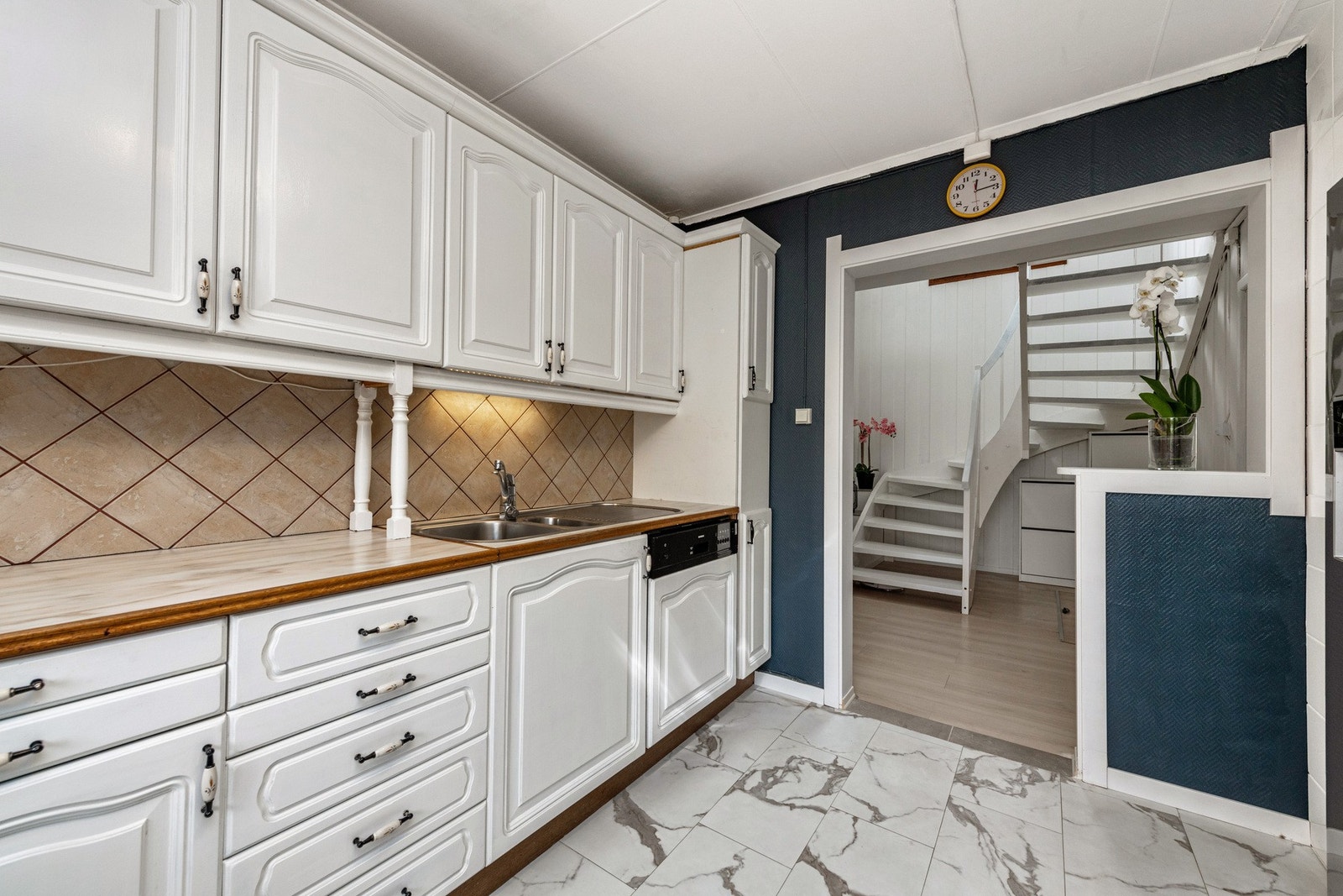 Fliser mellom benkeplate og overskap. Gulvet er flislagt. Stikkontakter over benk. Avsatt plass til frittstående kjøleskap med frysedel, platetopp og oppvaskmaskin er integrert i benken. Ventilator på veggen. Galleribilde