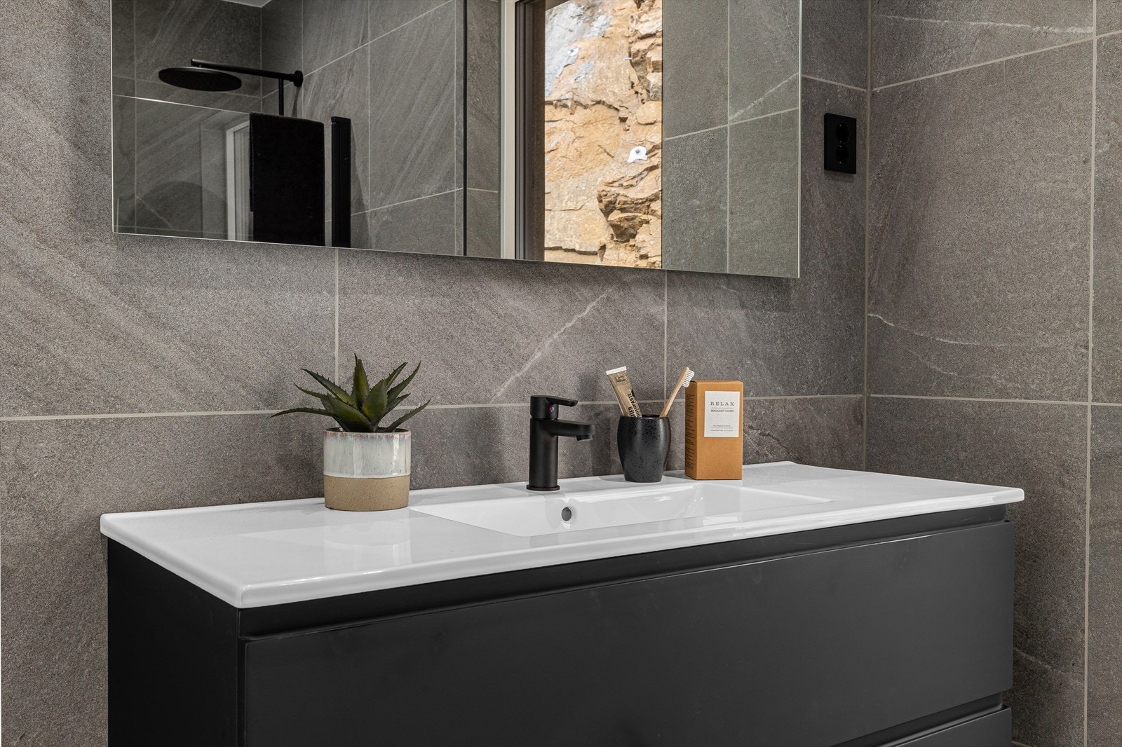 Badet har en mørk og moderne innredning med heldekkende servant på skuffeseksjon, speil, vegghengt toalett og dusjhjørne med foldedører i glass. Galleribilde