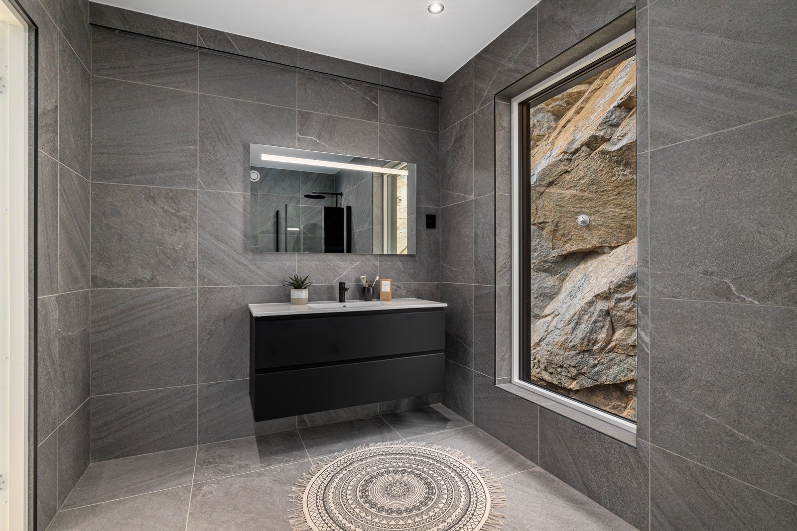 Badet er delikat og stilrent innredet med store, mørke 60x60 fliser, sorte detaljer, varme i gulv og downlights i himling. Galleribilde