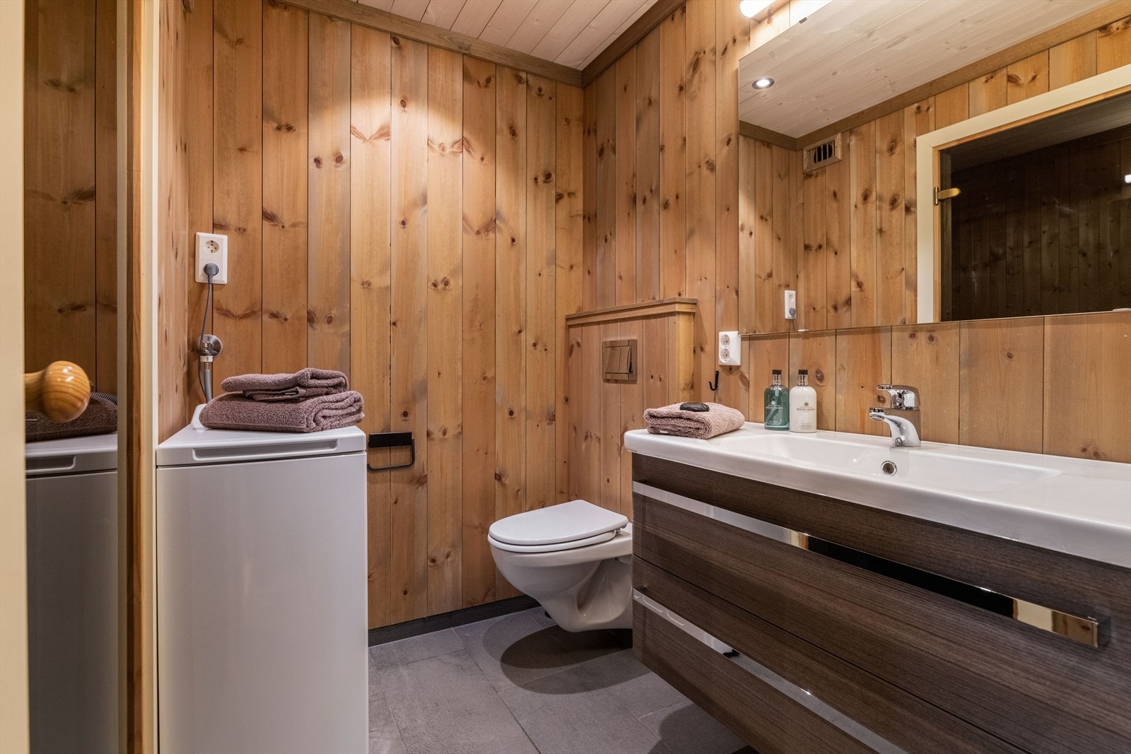 Badet inneholder vegghengt toalett, dusjhjørne med dusjdører og innredning med heldekkende hvit vask, underskuffer med slette fronter og speil. Det er mekanisk ventilasjon og opplegg for vaskemaskin. Galleribilde