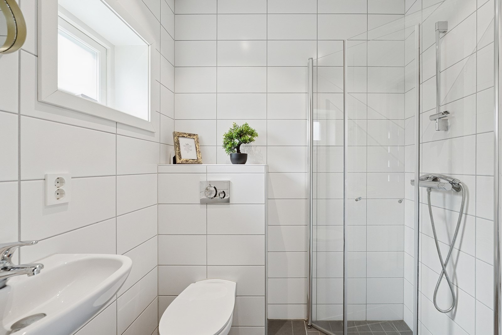 Baderommet har et stilrent uttrykk med flislagte vegger og gulv, samt elektriske varmekabler som gir en lun og behagelig komfort. Taket er malt og utstyrt med downlights som sørger for god belysning. Galleribilde