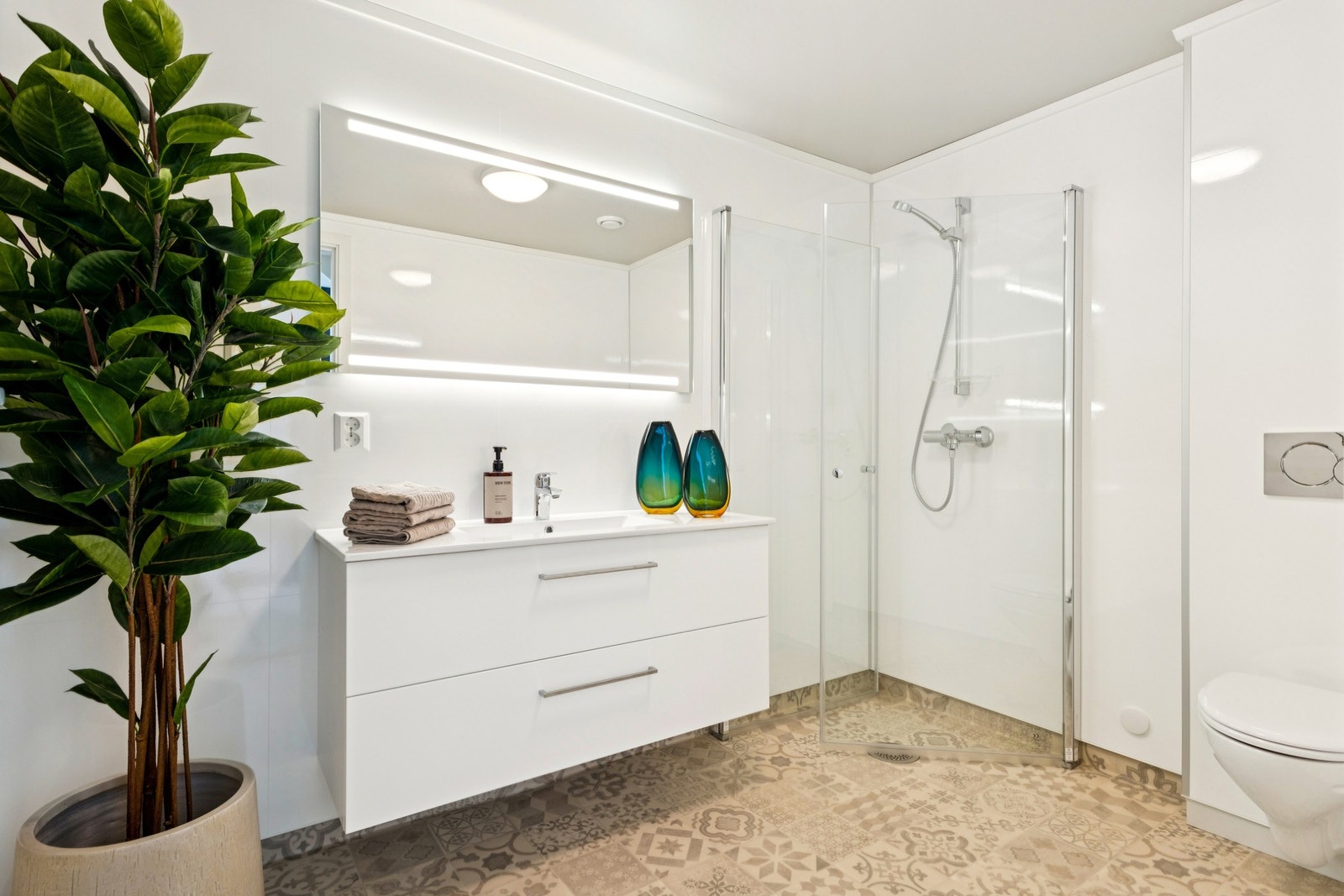 Badet er bygd med vinylgulv med varmekabler og våtromsplater på veggene. Innfellbart dusjhjørne, vegghengt toalett, opplegg for vaskemaskin og servant med speil og god belysning. Galleribilde