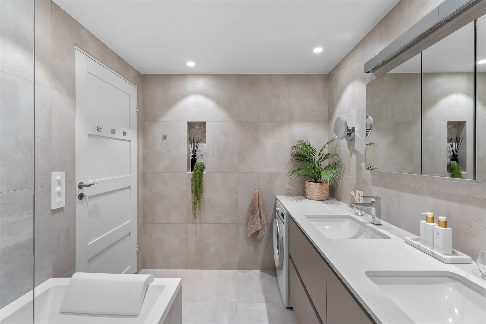 Dobbel servant med nedfelt vask i komposittbenk og speil med integrert belysning gir både stil og funksjonalitet. Spotter i taket og et innebygget badekar med dusj gir en behagelig atmosfære. Vegghengt toalett og opplegg for vaskemaskin. Galleribilde