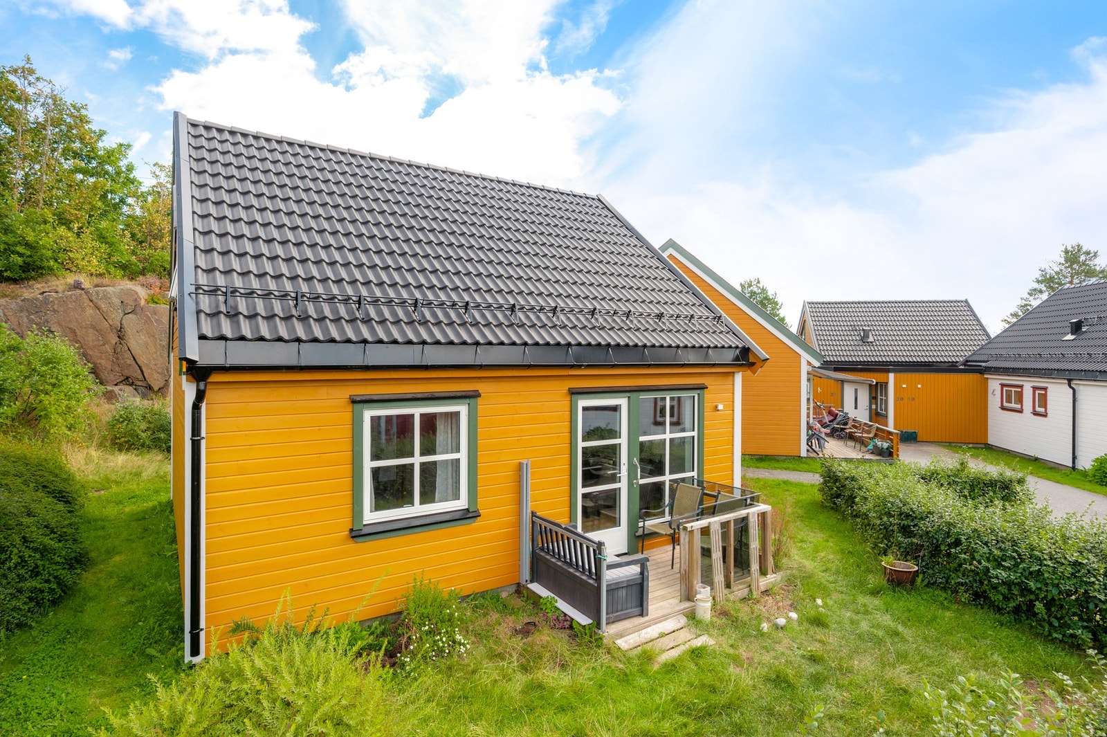 Vestvendt uteplass ut fra stuen med glassrekkverk og plen som omslutter boligen. Usjenert hagebakside - kveldssol. Galleribilde