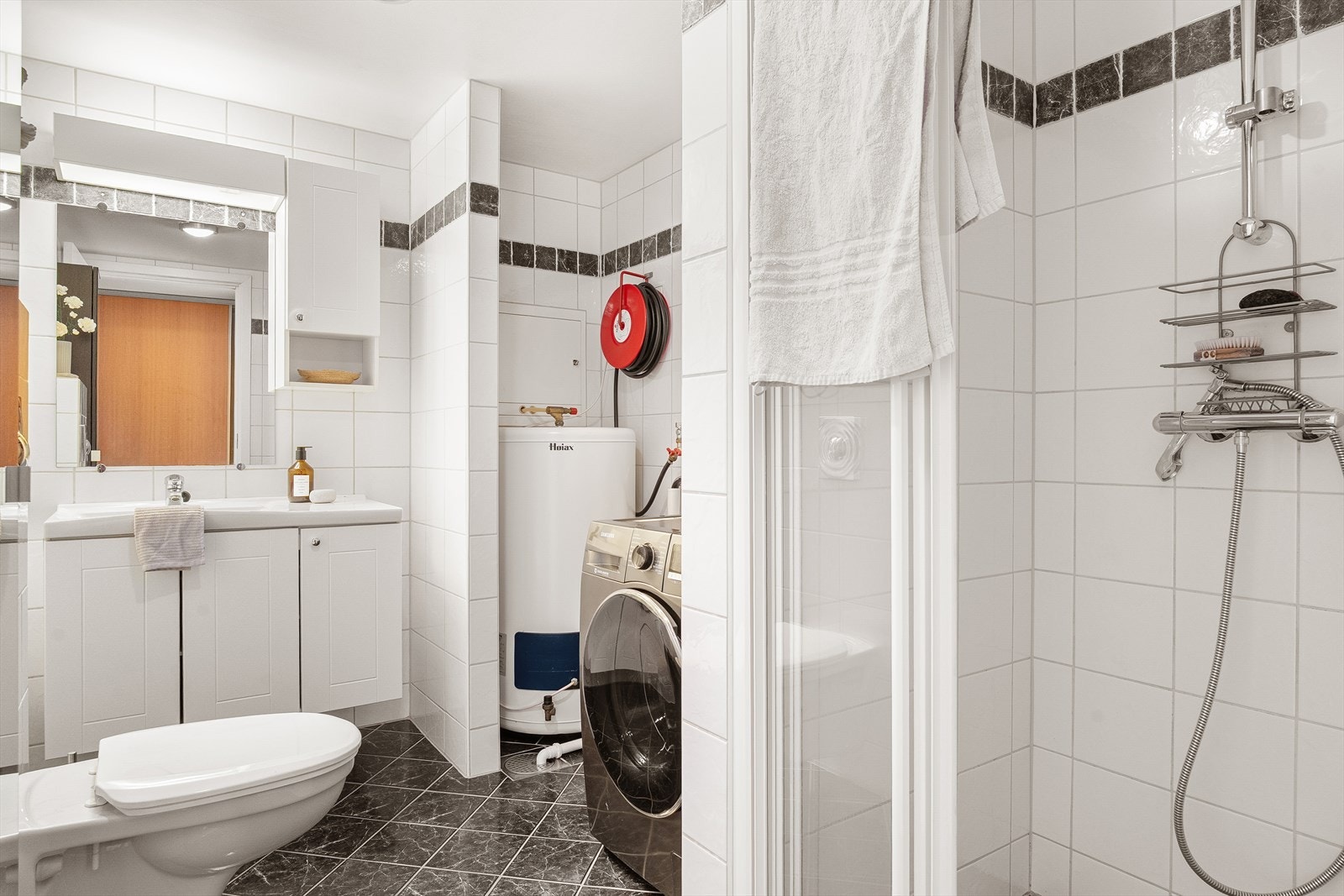 Helflist bad som er innredet med vegghengt toalett, stor dusjnisje, hvite baderomsmøbler og opplegg for vaskemaskin. Galleribilde