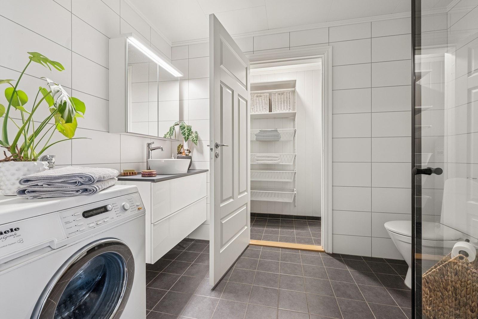 Pent flislagt baderom med toalett, dusjhjørne, servant og opplegg for vaskemaskin. Galleribilde