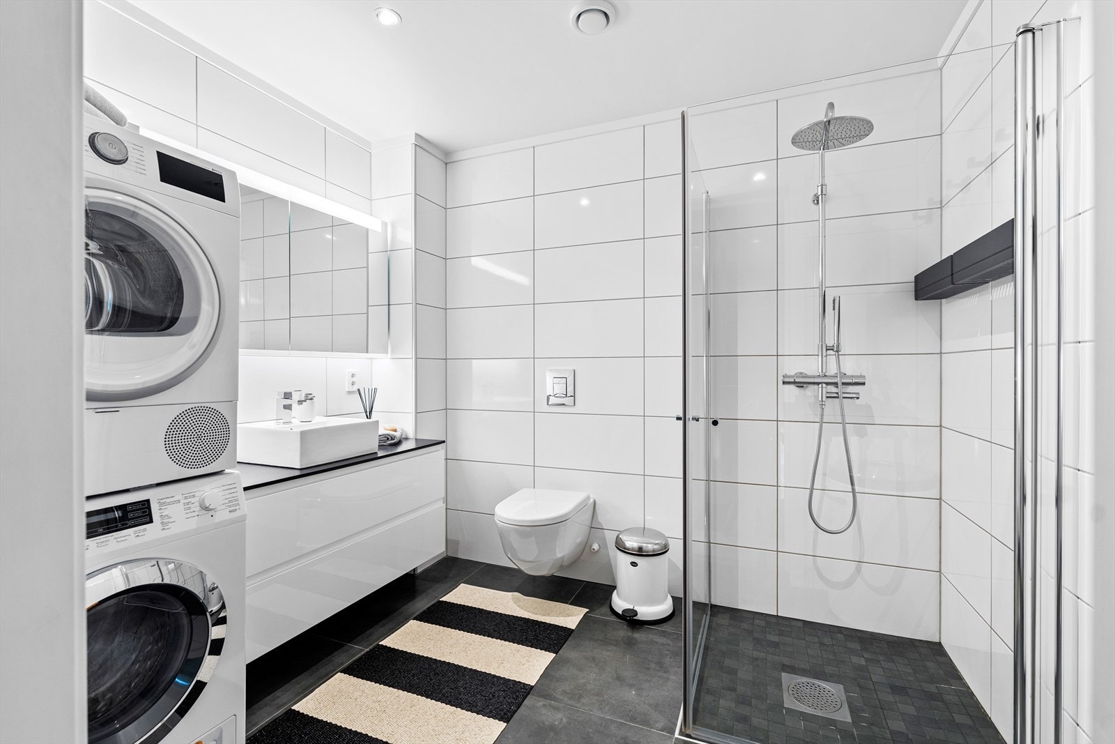 Stort flislagt bad med baderomsinnredning med glatte fronter, laminert benkeplate med påstående servant og speil over med tilhørende belysning. Galleribilde