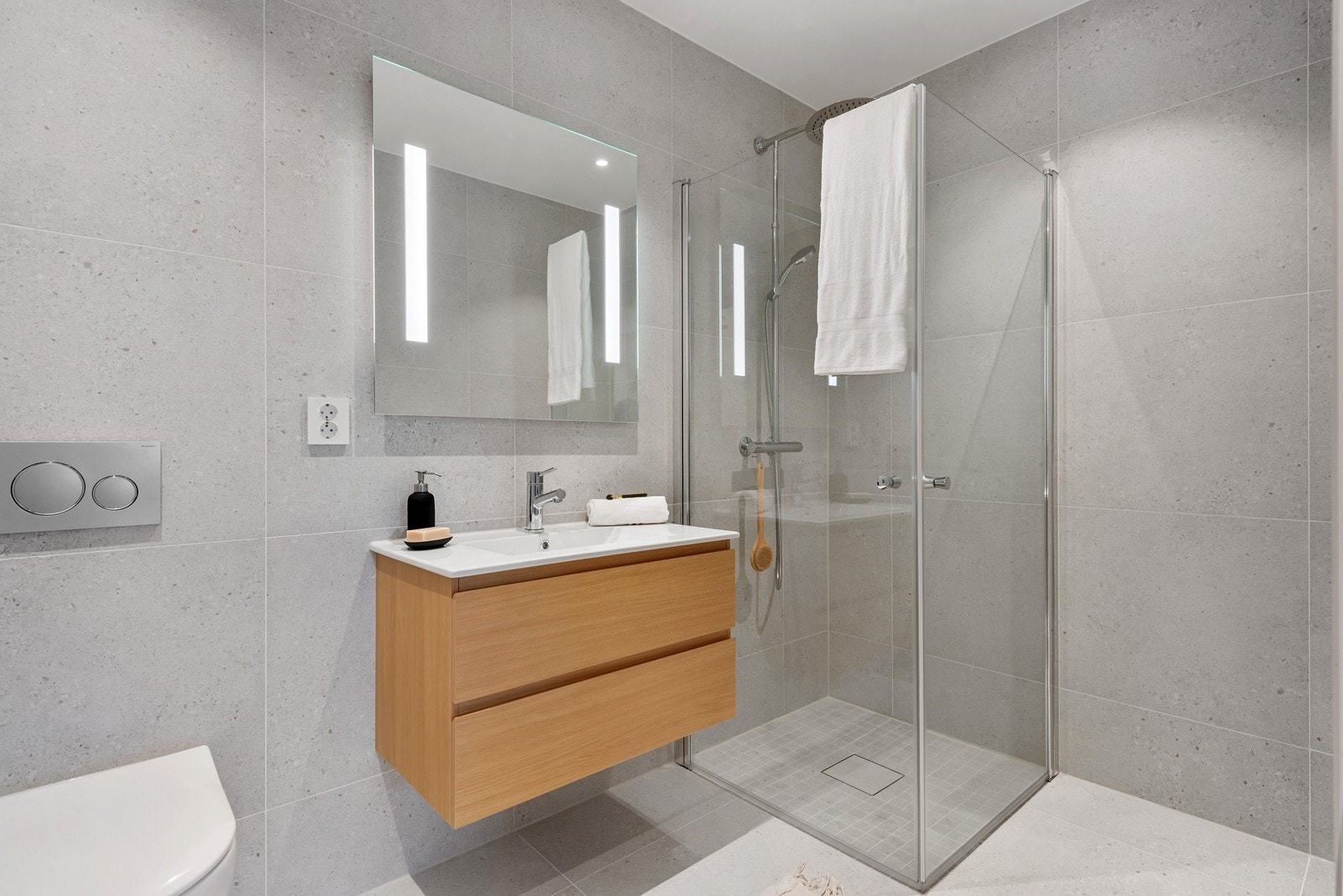 Badene er belagt med store fliser, og dusjhjørnet har svingbare glassdører samt hånd- og takdusj med armaturer i krom. Galleribilde