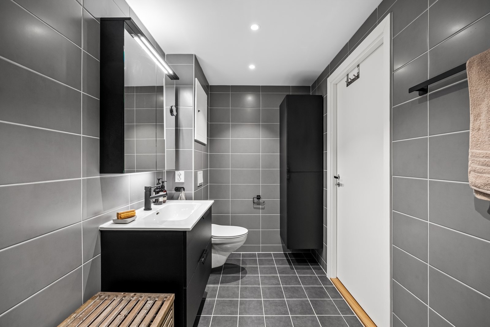 Badet er utstyrt med vegghengt toalett, dusjhjørne med glassdører, servantinnredning, speilskap med belysning og et praktisk høyskap for ekstra oppbevaring. Galleribilde