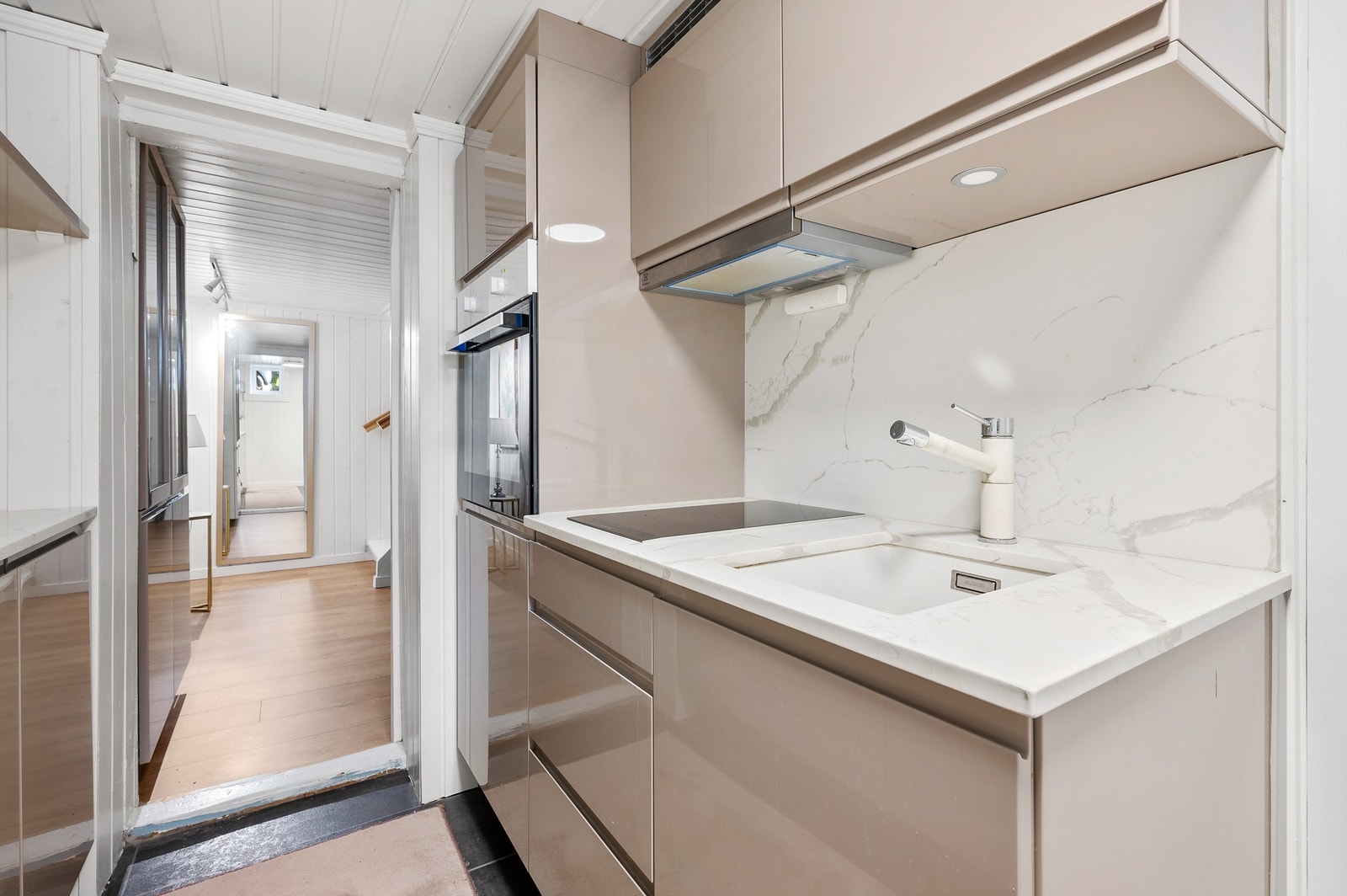 Innredningen har høyglansfronter med gripekant, hylle med belysning og hvitmarmorert benkeplate i stein med underlimt vaskekum i hvit kompositt. Kjøkkenet er utstyrt med stekeovn, smal induksjonstopp og kullfilterventilator. Galleribilde