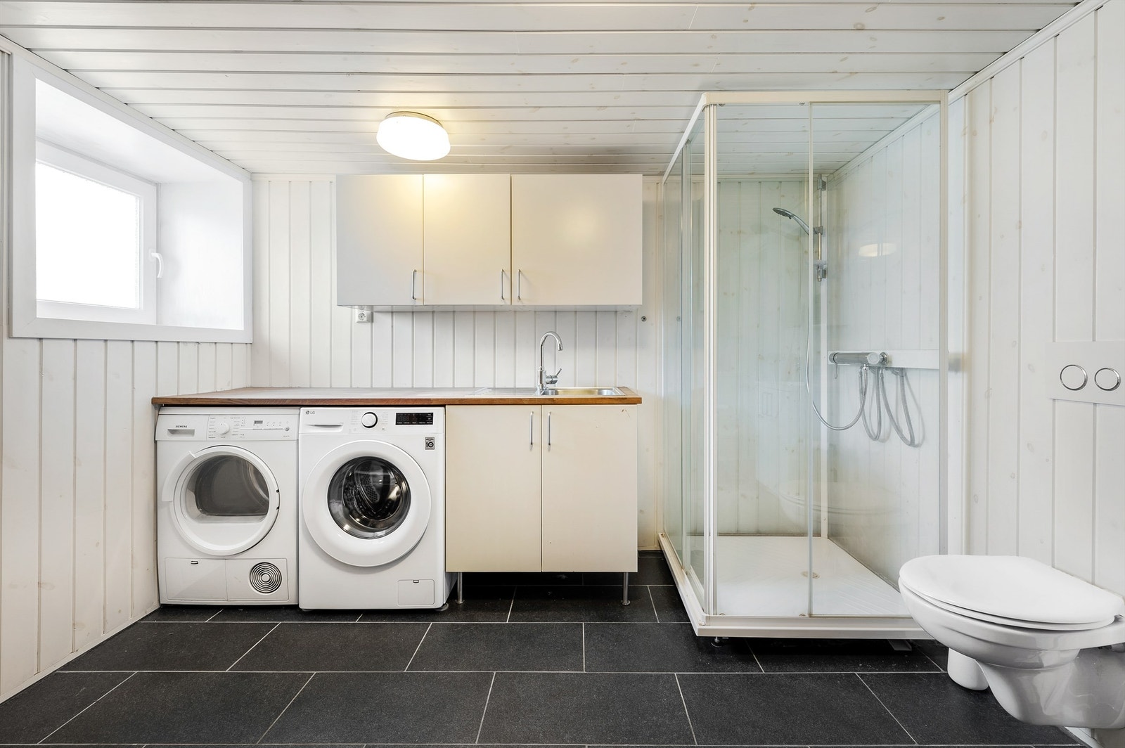 Innerst er det vaskeromsinnredning med lyse benkeskap, veggskap, benk og nedfelt vaskekum, samt nisje til vaskemaskin og tørketrommel under benken. Galleribilde