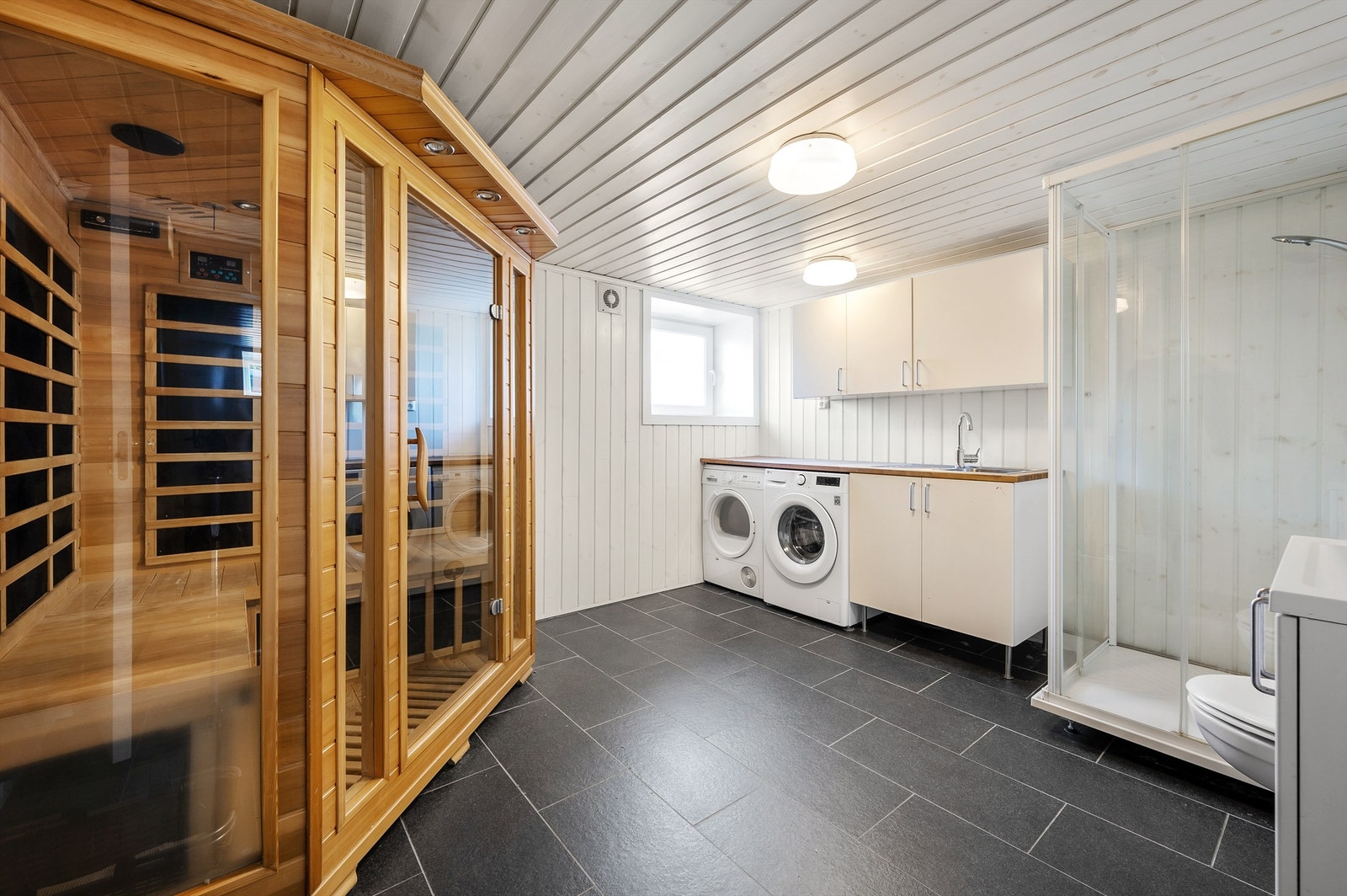 En stor, frittstående badstue med glassvegger, glassdør og plassbygde benker er plassert i hjørnet av rommet for ekstra velvære. Galleribilde