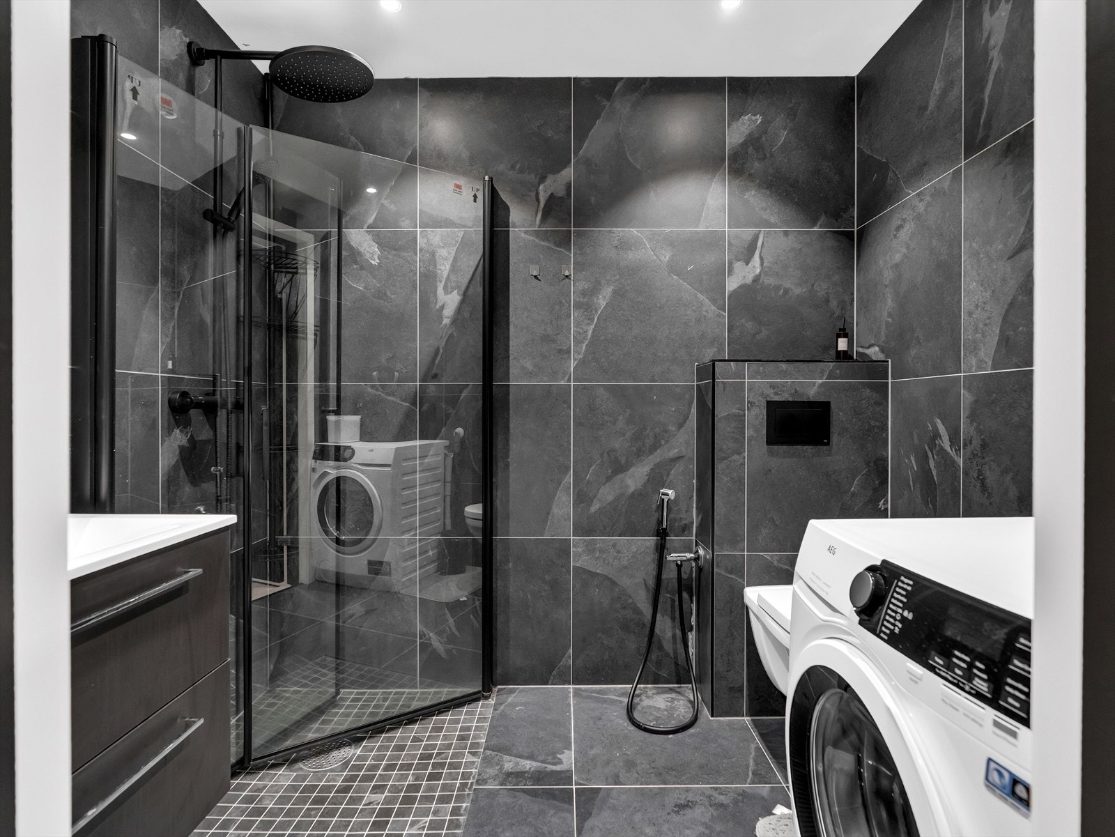 Delikat baderom med dusjhjørne med regnfallsdusj, vegghengt toalett, vegghengt innredning med servant og opplegg for vaskemaskin. Galleribilde