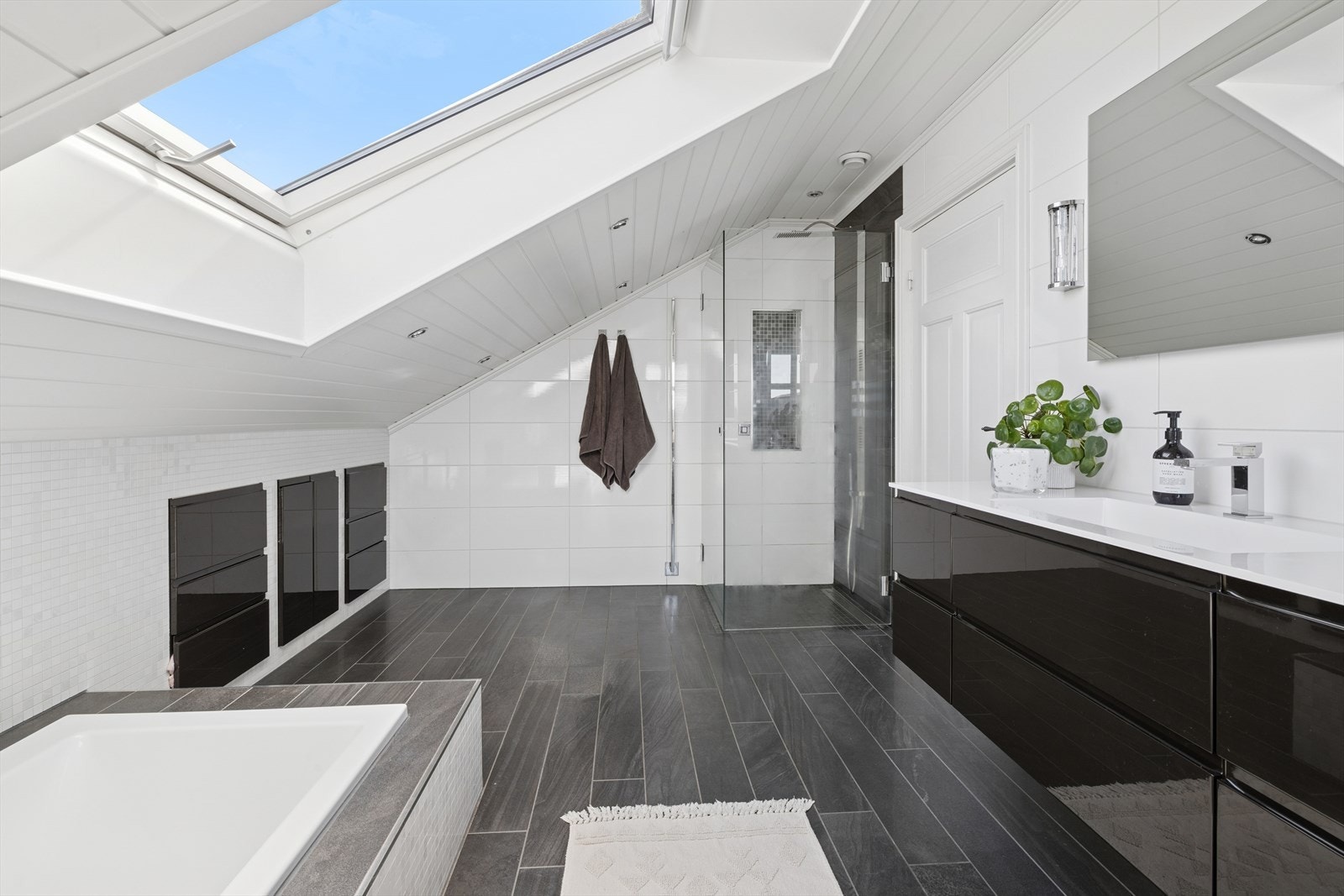 Badet har en baderomsinnredning med glatte fronter i høyglanset utførelse, speil og belysning over servant, servant i helstøpt plate, dusj med innfellbare vegger i herdet glass, badekar nedsenket i gulv og veggfestet klosett med innebygget sisterne. Galleribilde