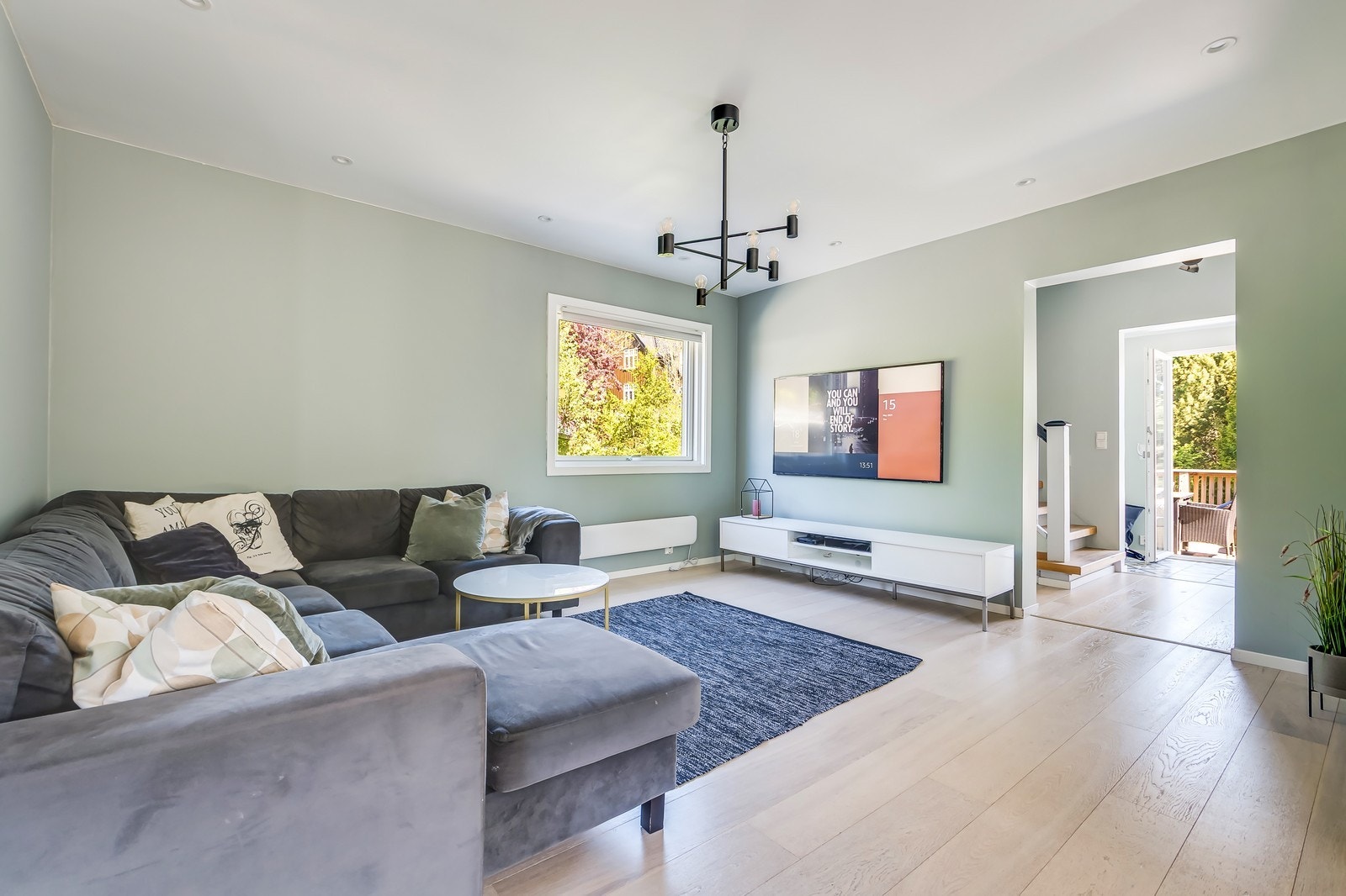 Stue med grønnmalte vegger. Plass til stor grå hjørnesofa, stuebord, TV benk og vegghengt TV. Galleribilde