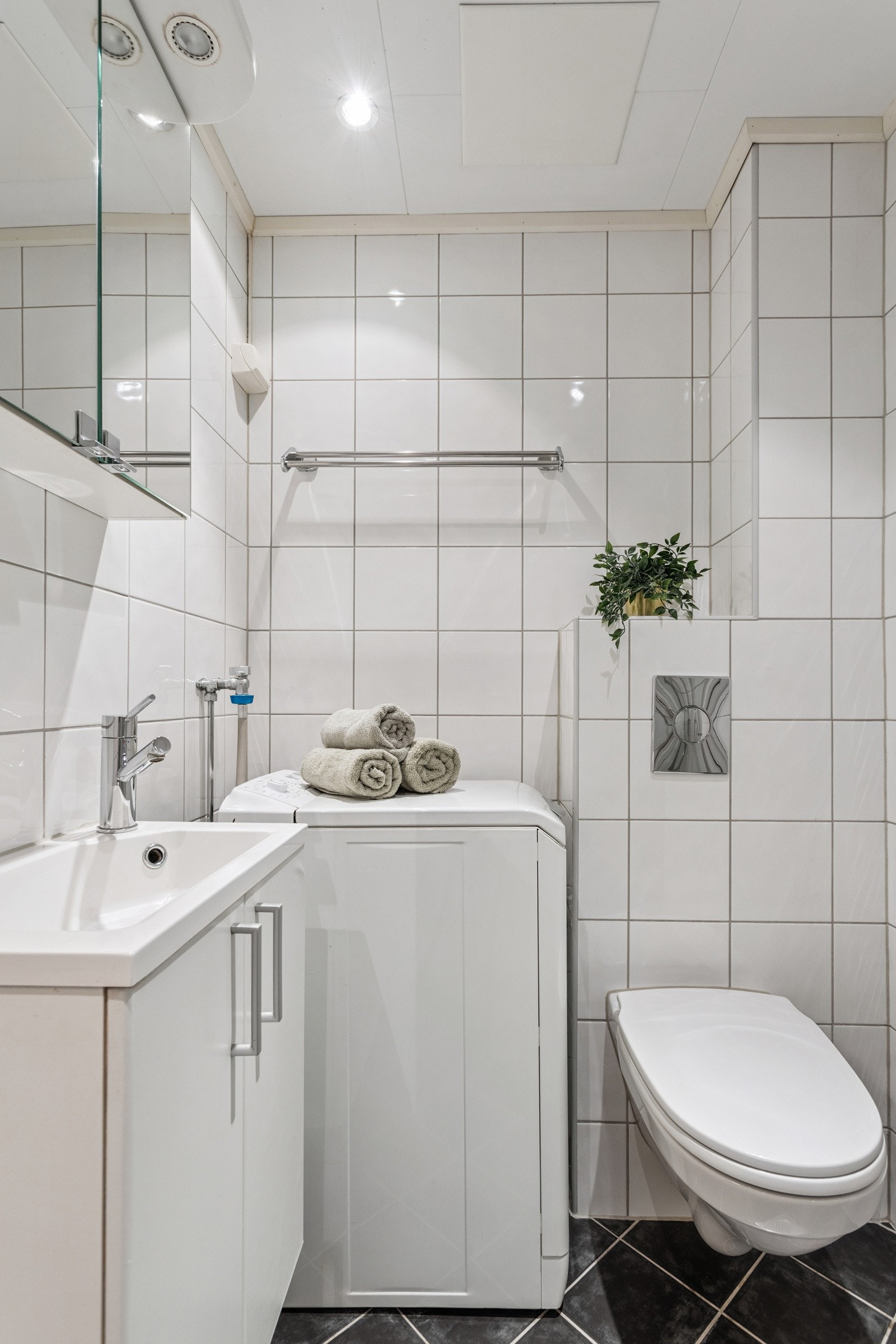 Flislagt bad med dusj med inn-/utadslående dusjvegger, servant med underskap, samt speilskap. Veggmontert klosett og opplegg for vaskemaskin (vaskemaskin medfølger). Galleribilde