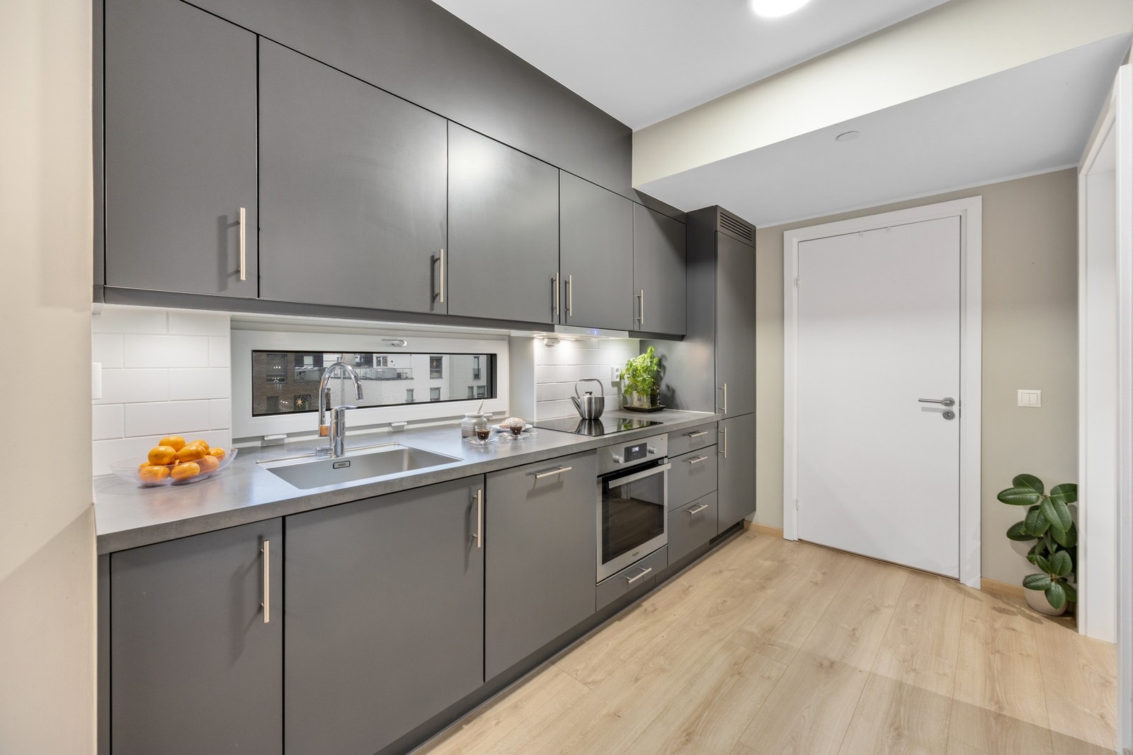 Kjøkkeninnredningen består av grå slette fronter og laminat benkeplate med nedfelt oppvaskkum i rustfritt stål. Det er belysning under oversskap og fliser på vegg over benkeskap. Av integrerte hvitevarer medfølger stekeovn, platetopp, oppvaskmaskin og kjø Galleribilde