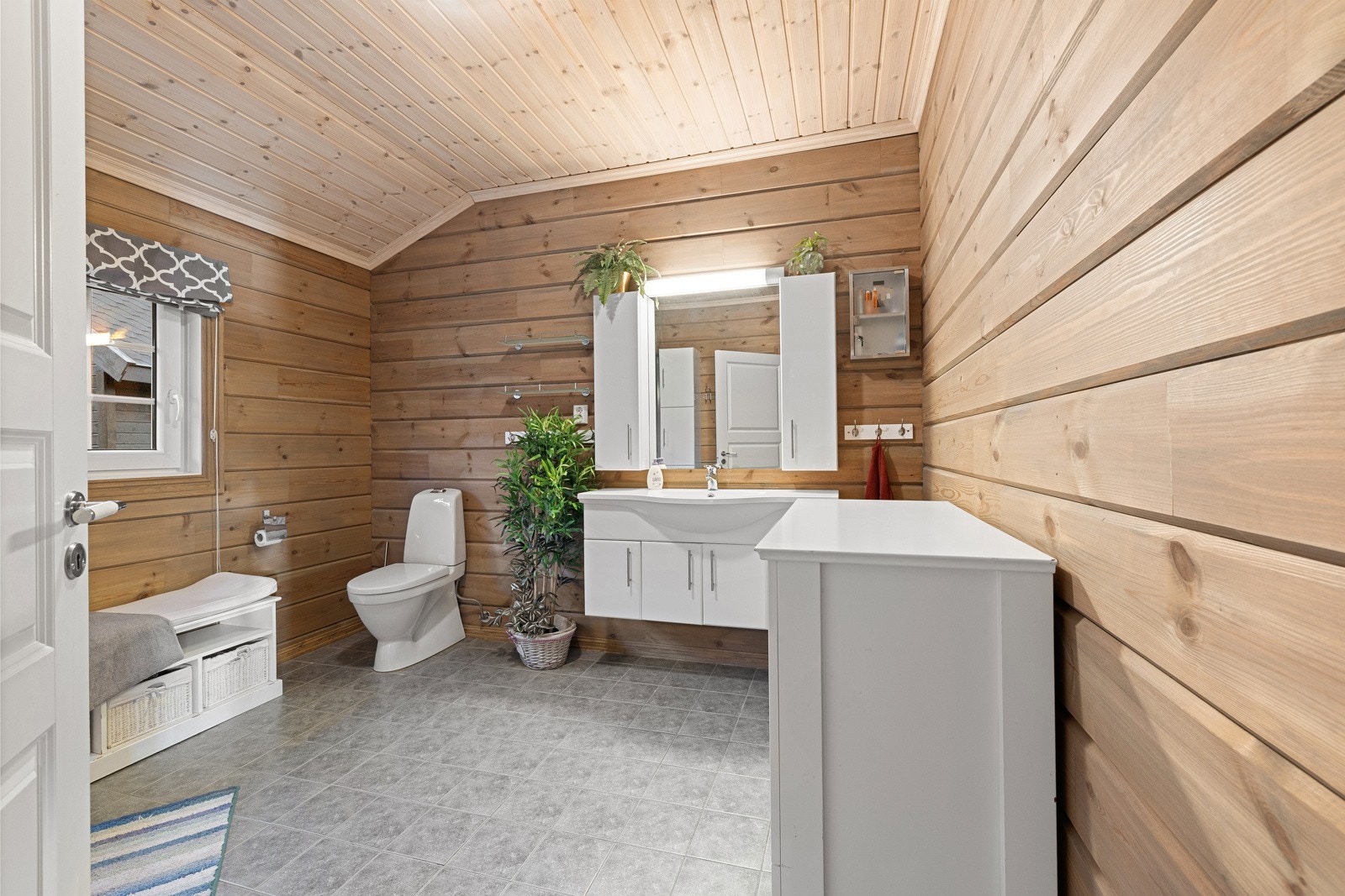 Romslig bad innredet med dusjkabinett, gulvmontert toalett og baderomsinnredning med nedsenket servant og speil. Gulvet er flislagt med elektriske varmekabler, mens vegger er kledd med tømmer og trepanel. Galleribilde