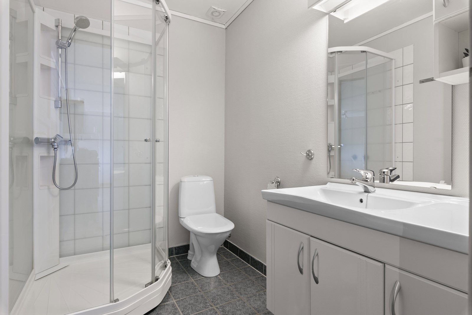 Badet har flislagt gulv med varmekabler og tapet på vegg. Badet inneholder servant med skap, wc og dusjkabinett. Galleribilde