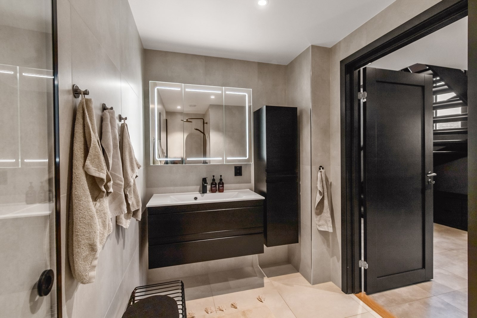 Nede er badet innredet med store, delikate fliser; beige på gulvet og grå på veggene. Varmekabler er installert, og badet har downlights og vegghengt toalett. Galleribilde