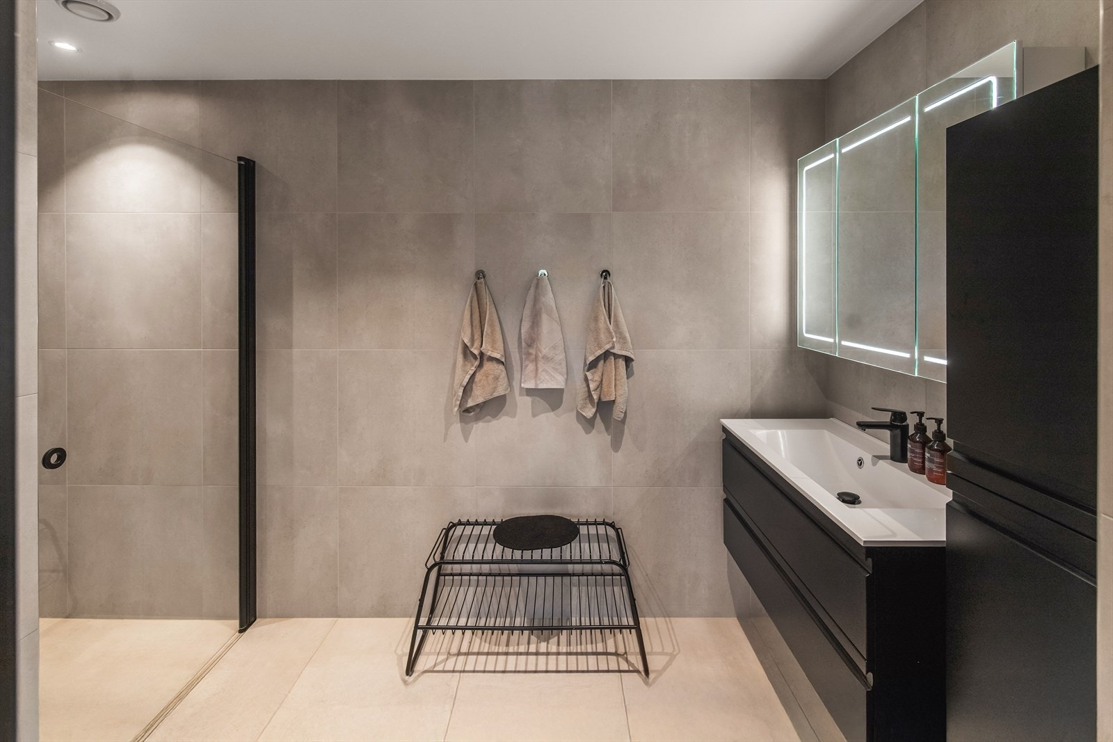 Et dusjhjørne er utstyrt med sort regnfallsdusj og svingbar glassdør i sort innramming. Baderomsinnredningen består av servant med sorte skuffer, et matchende høyskap og et stort speilskap med integrert belysning. Galleribilde