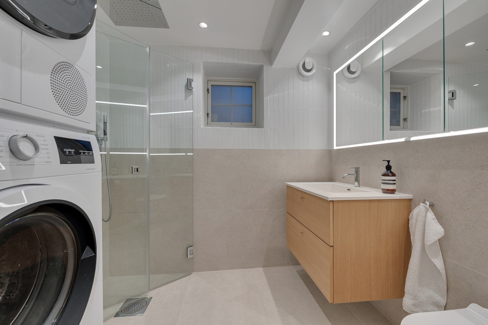 Nydelig bad fra 2024 vegghengt wc, servant med skuffer under, speilskaper med lys rundt og takdusj i hjørne med innad slående dører. Det er opplegg for vaskemaskin og tørketrommel. Galleribilde