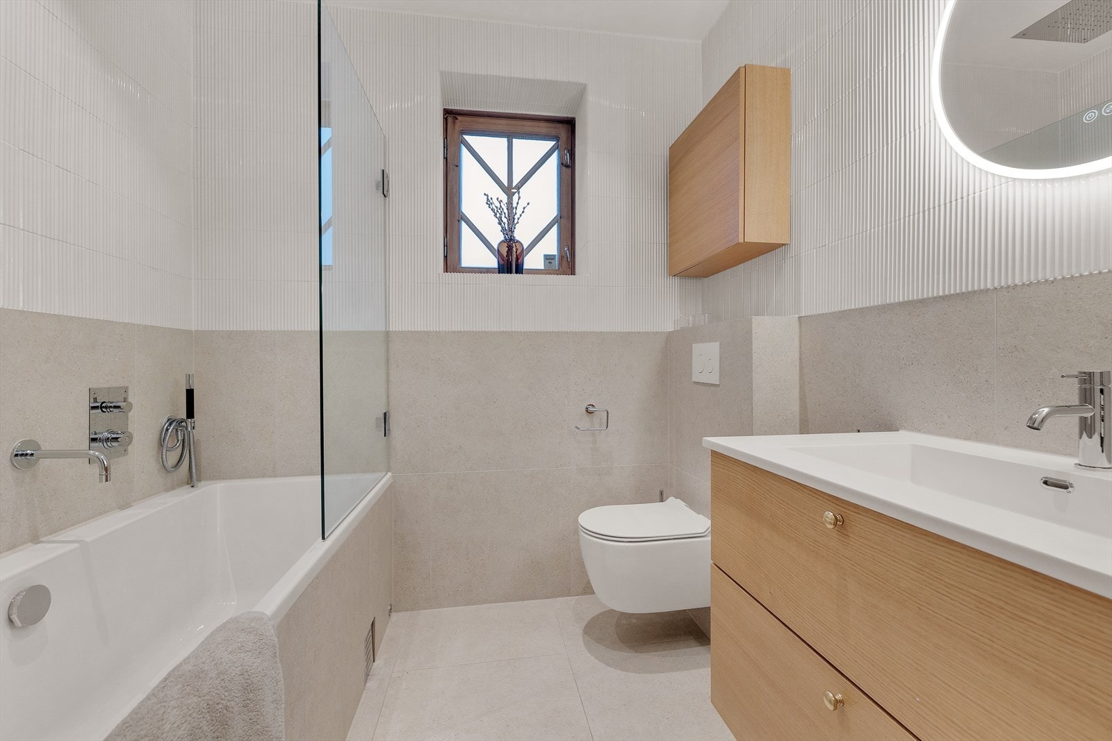 Pent og moderne bad fra 2024 med vegghengt wc, servant med skuffer under, speil med lys rundt og takdusj i innmurt badekar. Galleribilde