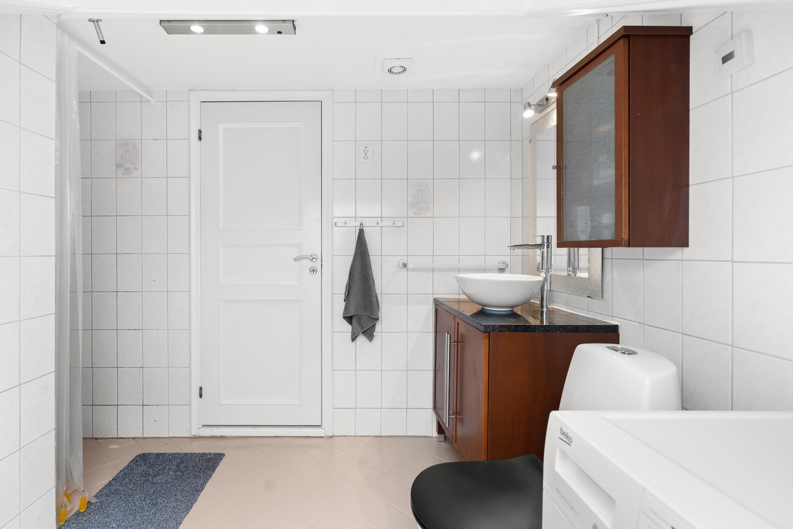 Rommet er utstyrt med dusj på gulv, gulvstående klosett, servant og benkeskap med profilerte fronter. I tillegg har badet opplegg for vaskemaskin. Galleribilde