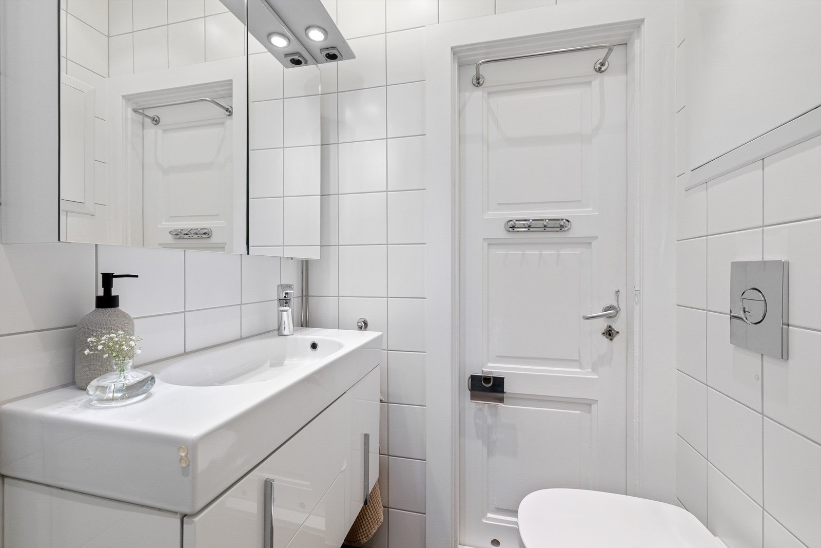 Badet er innredet med dusjhjørne med innfellbare glassdører og hånd- og regndusj, veggmontert toalett og baderomsinnredning med en bredde på 80 cm med skuffer, overlimt servant med ettgreps armatur fra Oras og speilskap med spotter. Galleribilde