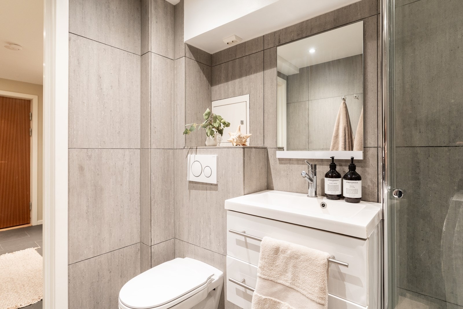 Dusjen er plassert i hjørnet og har en nedfelt dusjnisje med mosaikkfliser samt håndholdt dusjarmatur. Videre er badet utstyrt med vegghengt toalett, opplegg for oppvaskmaskin og elektrisk styrt vifte i taket. Galleribilde