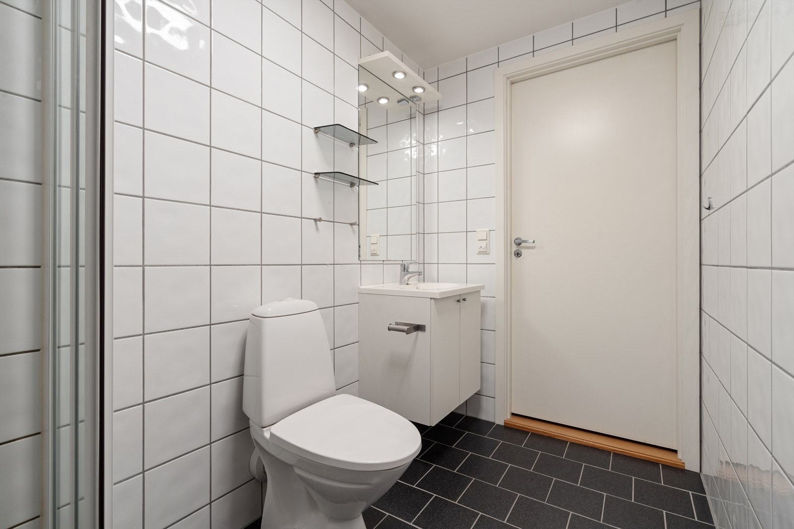 Badet er innredet med gulvmontert toalett, opplegg for vaskemaskin, dusjnisje med dusjgarnityr på vegg, og servant. Galleribilde