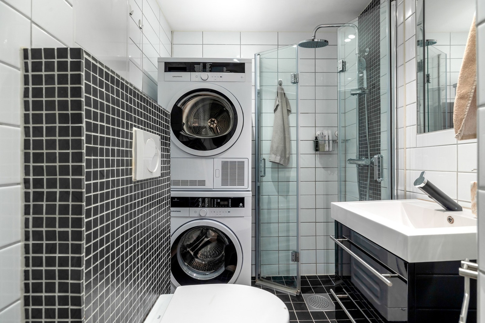 Badet er flislagt med lyse vegger og mørkt gulv. Rommet er innredet med dusjhjørne med glassvegger og regnfalldusj, vegghengt toalett samt servant med oppbevaringsløsning i undersuffer. Galleribilde