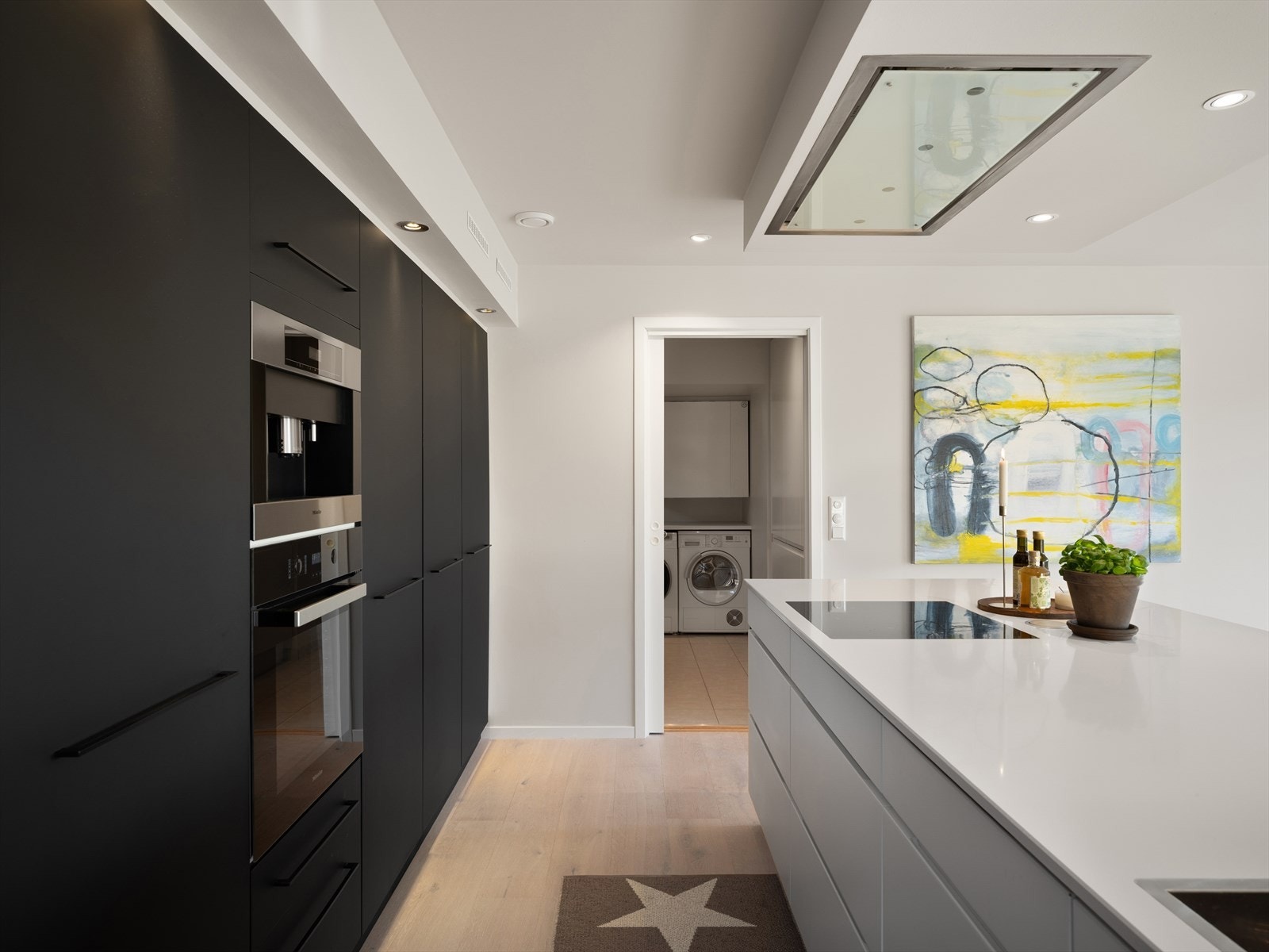 Siemens koketopp, integrert Miele kaffemaskin. Miele stekeovn og ASKO oppvaskmaskin (2025) Pene kontraster mellom sorte fronter på høyskap (2019) og hvit kjøkkenøy. Benkeplate av Silestone, og Eiko distante ventilator over kjøkkenøy. Galleribilde