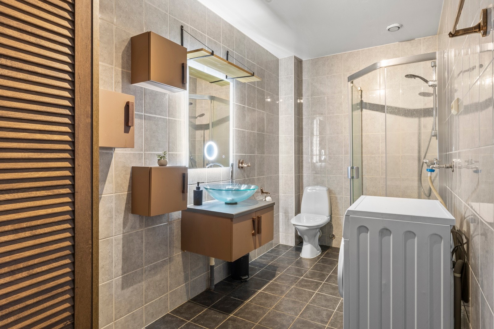 Stort bad/vaskerom med fliser å gulv og vegger, stilig baderomsmøbel med servant, dusjhjørne og opplegg for vaskemaskin. Galleribilde