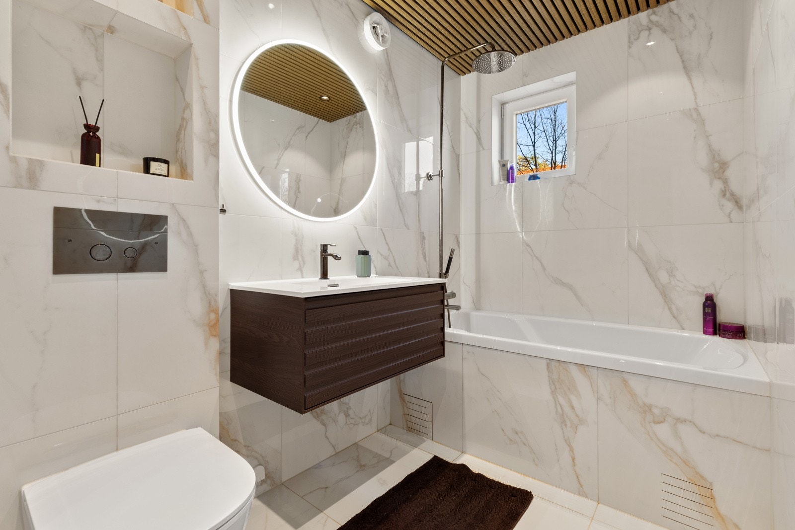 Baderommet har flotte fliser på gulv og vegger, deilig gulvvarme og stilige trespiler med innfelte downlights i himlingen. Det er også et lite vindu som var nytt i 2023 og mekanisk avtrekksvifte i vegg. Galleribilde