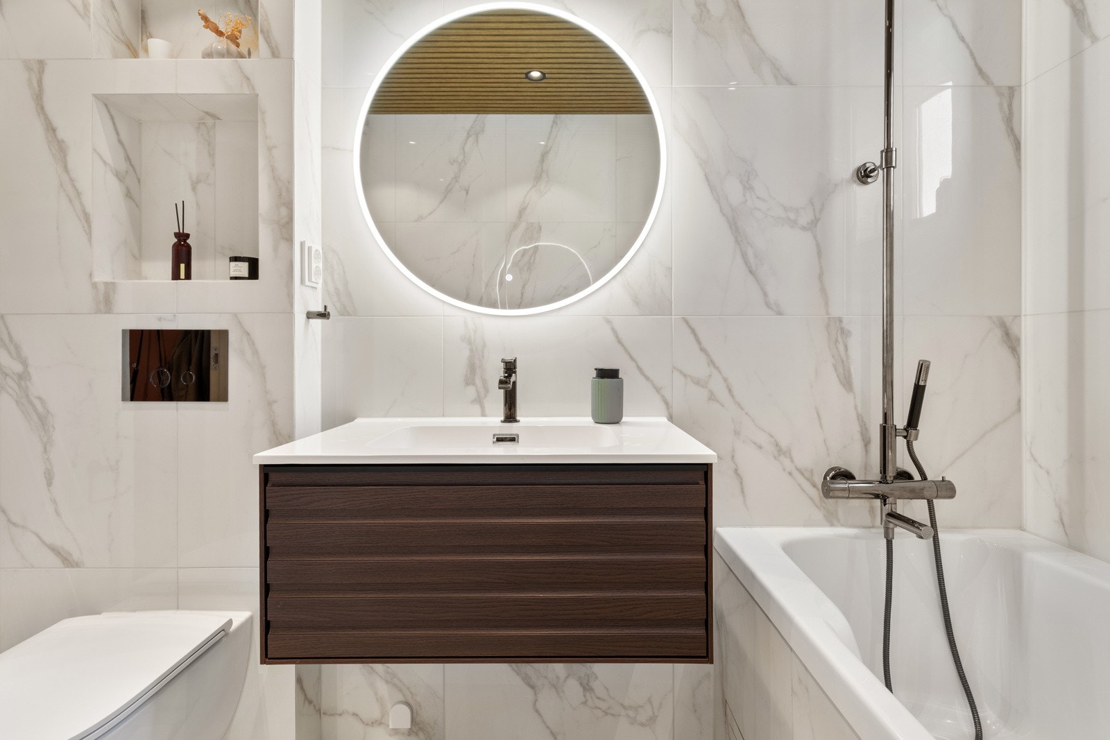 Flott baderomsinnredning med skuff og heldekkende servant, rundt speil med integrert belysning over. Innerst i rommet er det innmurt badekar og vegghengt dusj med både hånd- og regnfallsdusj. Galleribilde