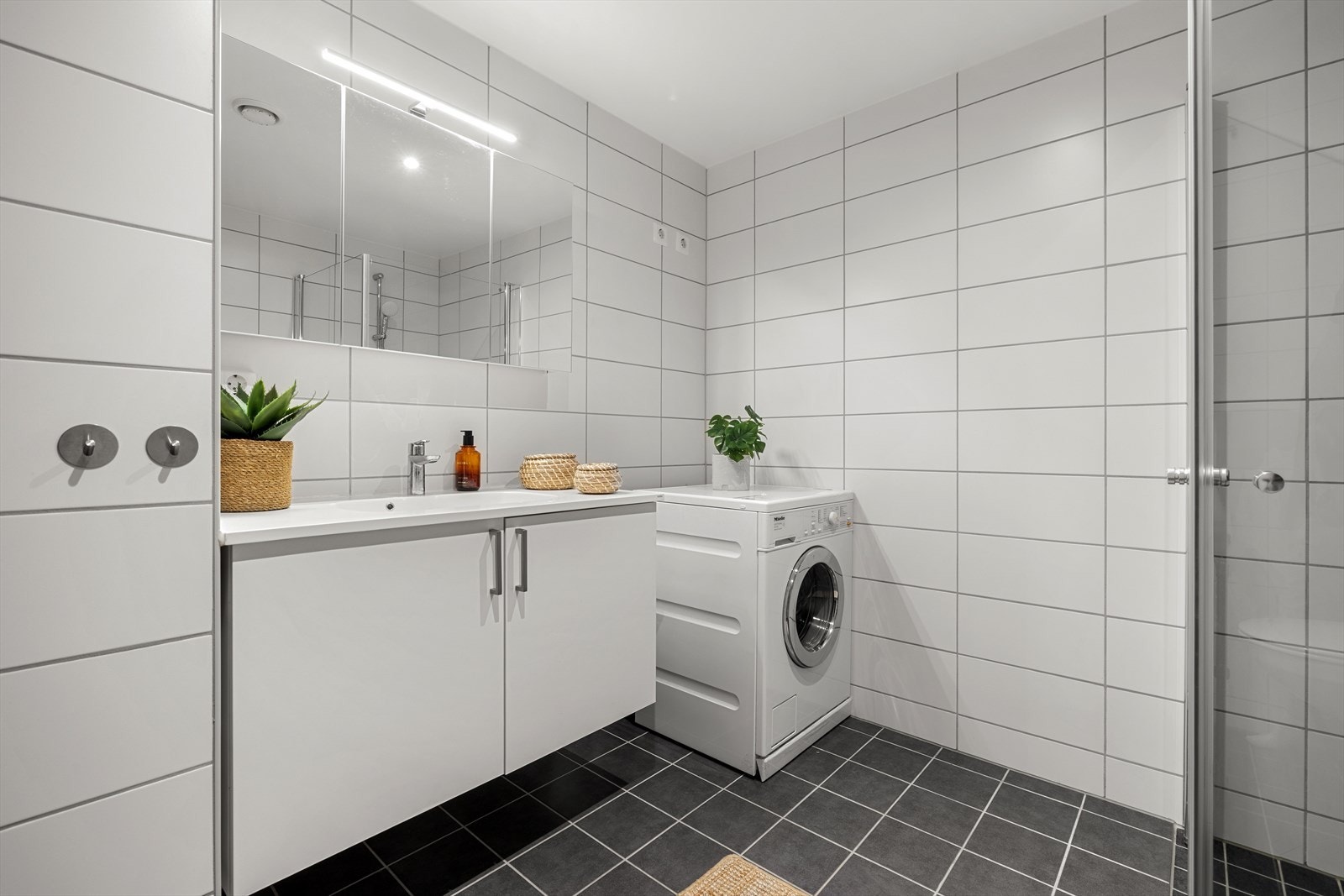 Videre er badet utstyrt med innredning bestående av nedfelt servant med underskap og 1-greps blandebatteri, speil, dusjhjørne med glassdører, vegghengt toalett og opplegg for vaskemaskin. Galleribilde