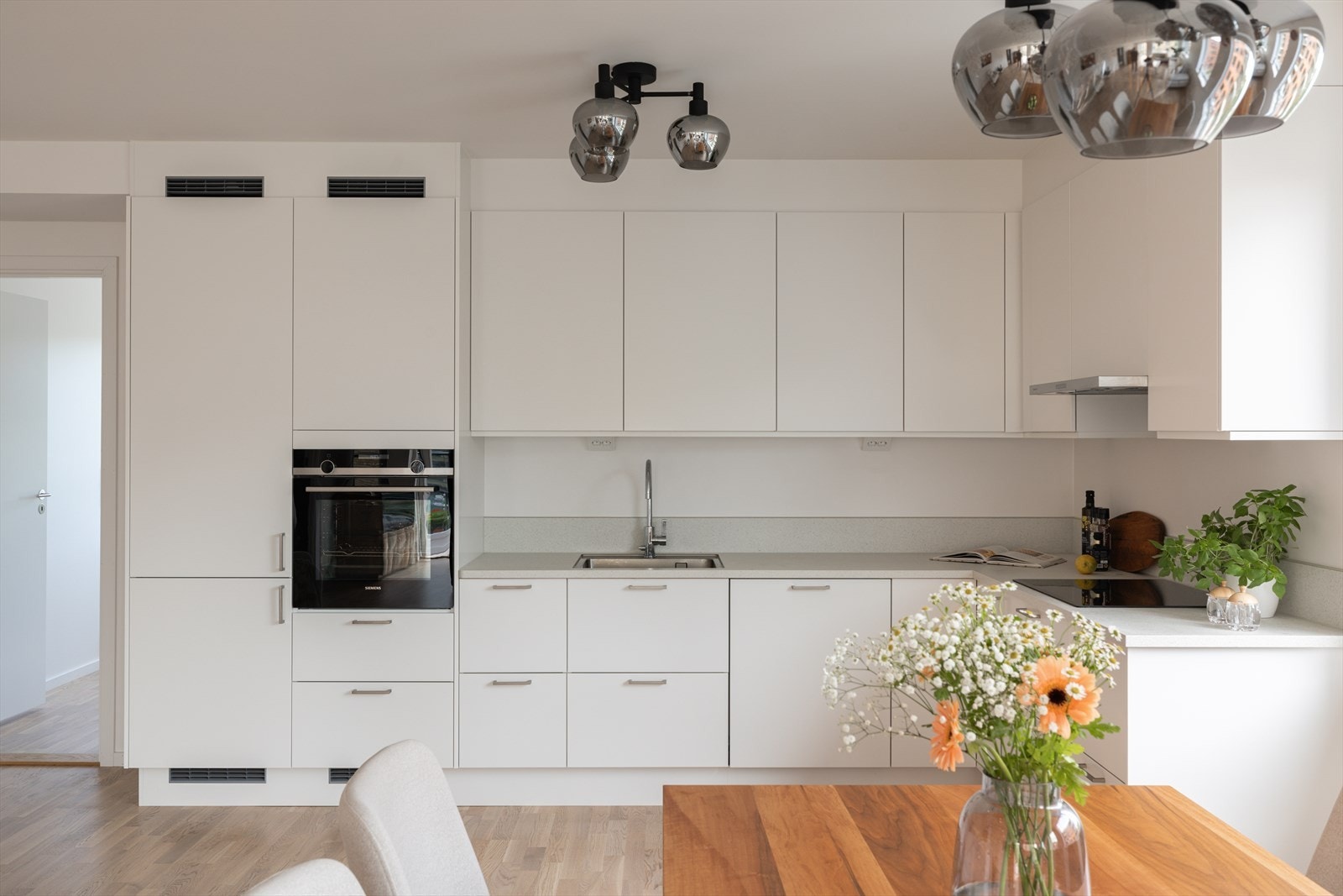 Innredningen har takoppforede skap, hvite, glatte fronter, og en lys benkeplate i laminat med lav backsplash mot benk og en vaskekum i stål. Galleribilde