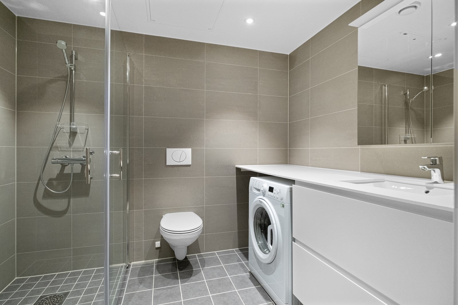 Stort baderom med romslig utførelse og innredning. Badet inneholder dusjhjørne, vegghengt toalett, rom og opplegg til vaskemaskin i hjørne, servant, overhengt skap med speil Galleribilde