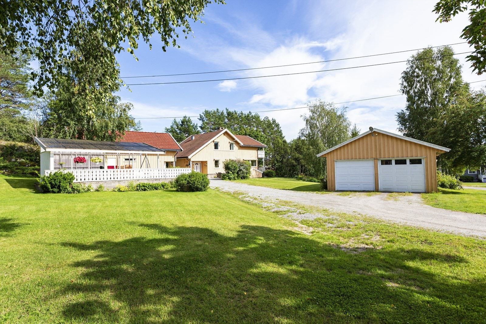 Sommerbilde. En eiendom bestående av stort våningshus, garasje/kontor, stabbur og en hagestue med tilliggende terrasse. Galleribilde
