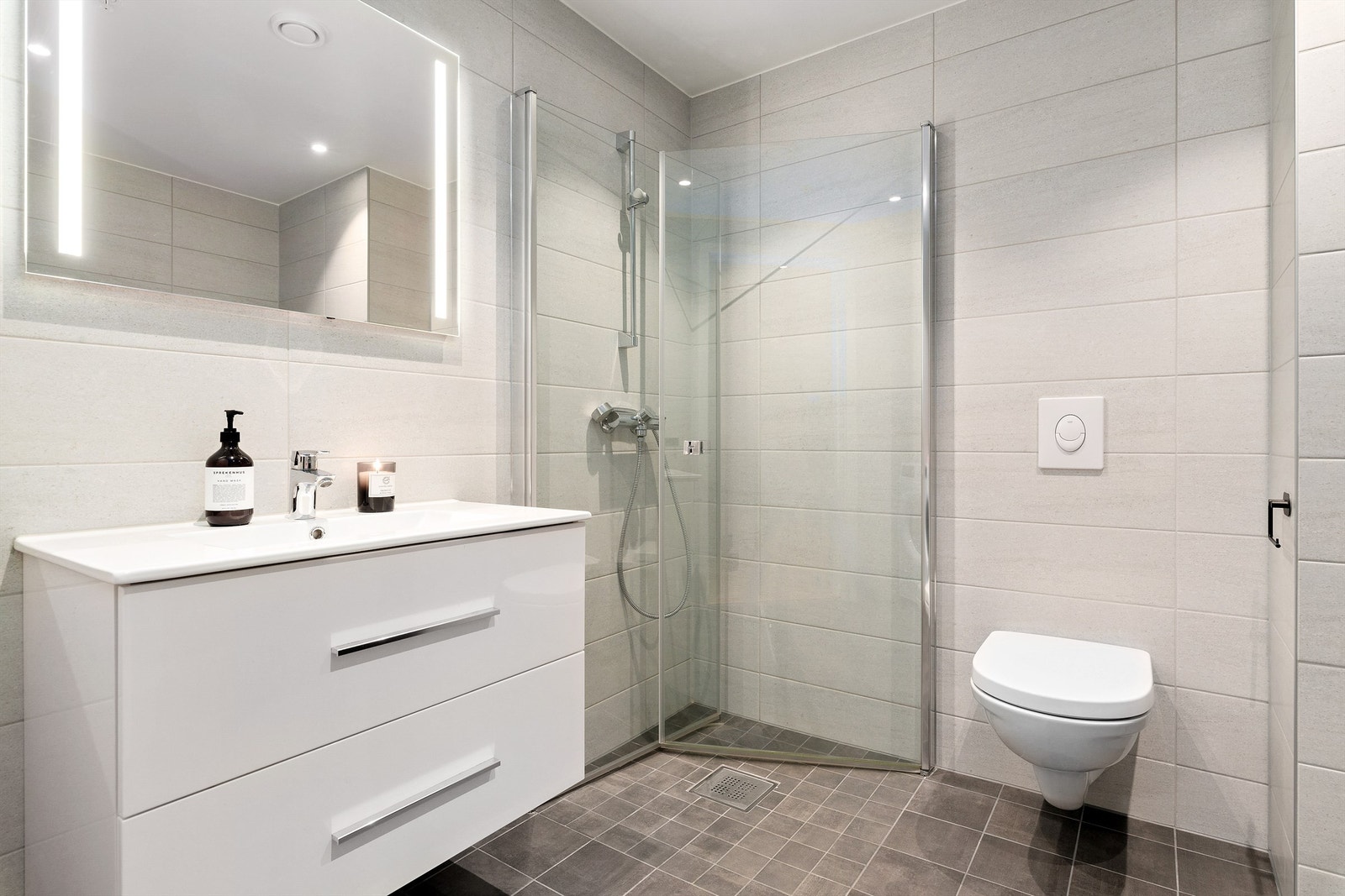 Bad/wc med fliser på gulv og vegger. Malte flater i himling og spotter i tak. Vegghengt servantinnredning og vegghengt dusjarmatur. Speil med sidelys og stikkontakt over servant Galleribilde