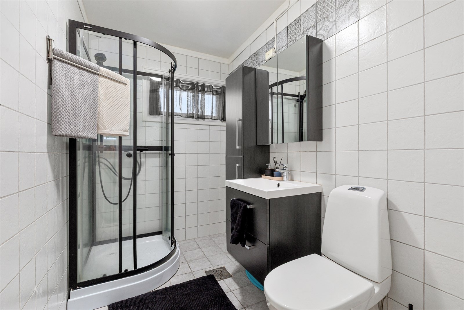 Bad med fliser på gulv og vegger og kvitmalt slett himling. Badet er utstyrt med dusjkabinett, toalett og servant med innredning. Galleribilde