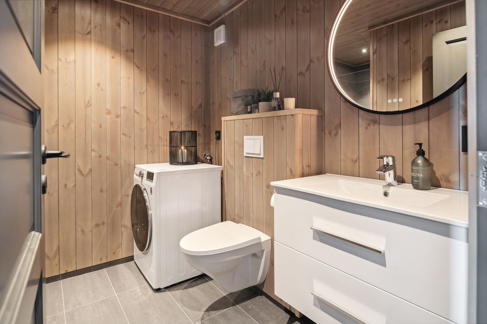 Baderom med delvis panelkledde vegger og gulvfliser med gulvvarme. Vegghengt wc, helservant på skuffeinnredning, speil m/belysning og downlights i himling. Det er opplegg for vaskemaskin. Galleribilde