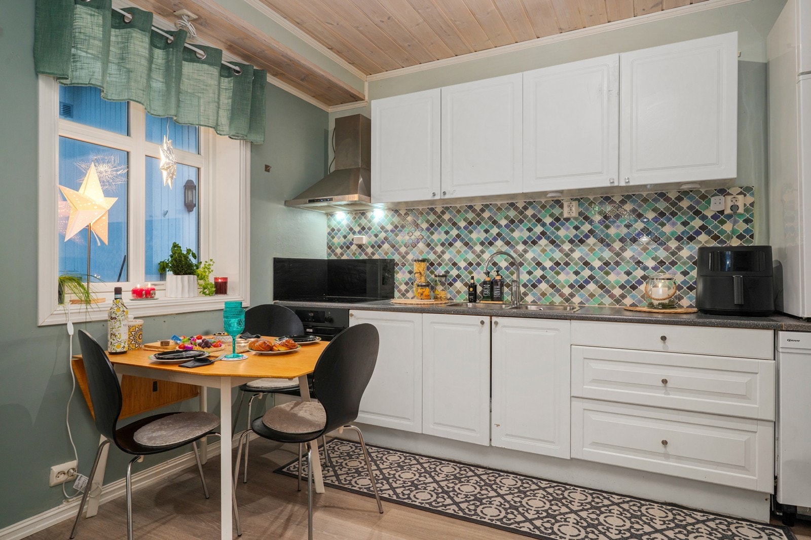 Hvite fronter og fargerik flislagt bakvegg gir kjøkkenet et friskt uttrykk. Det er plass til et lite spisebord ved vinduet. Galleribilde