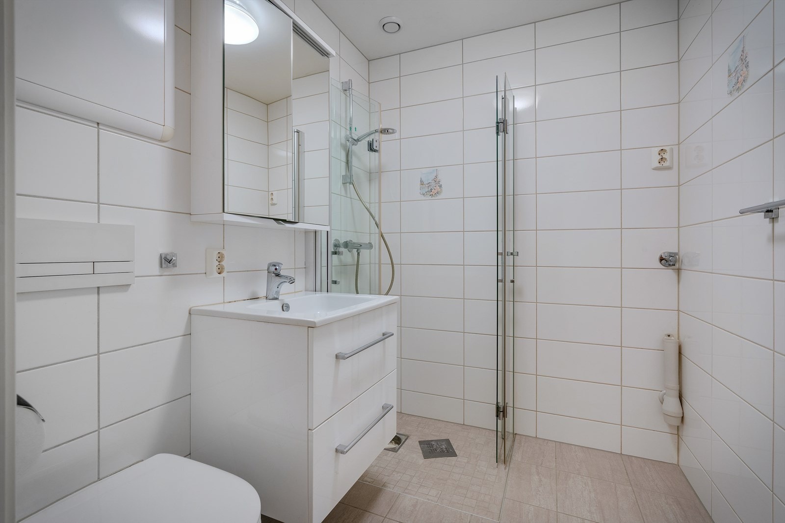 Baderommet er flislagt med foldedører i dusjen, veggtoalett og baderomsinnredning med heldekkende servant og speil Galleribilde