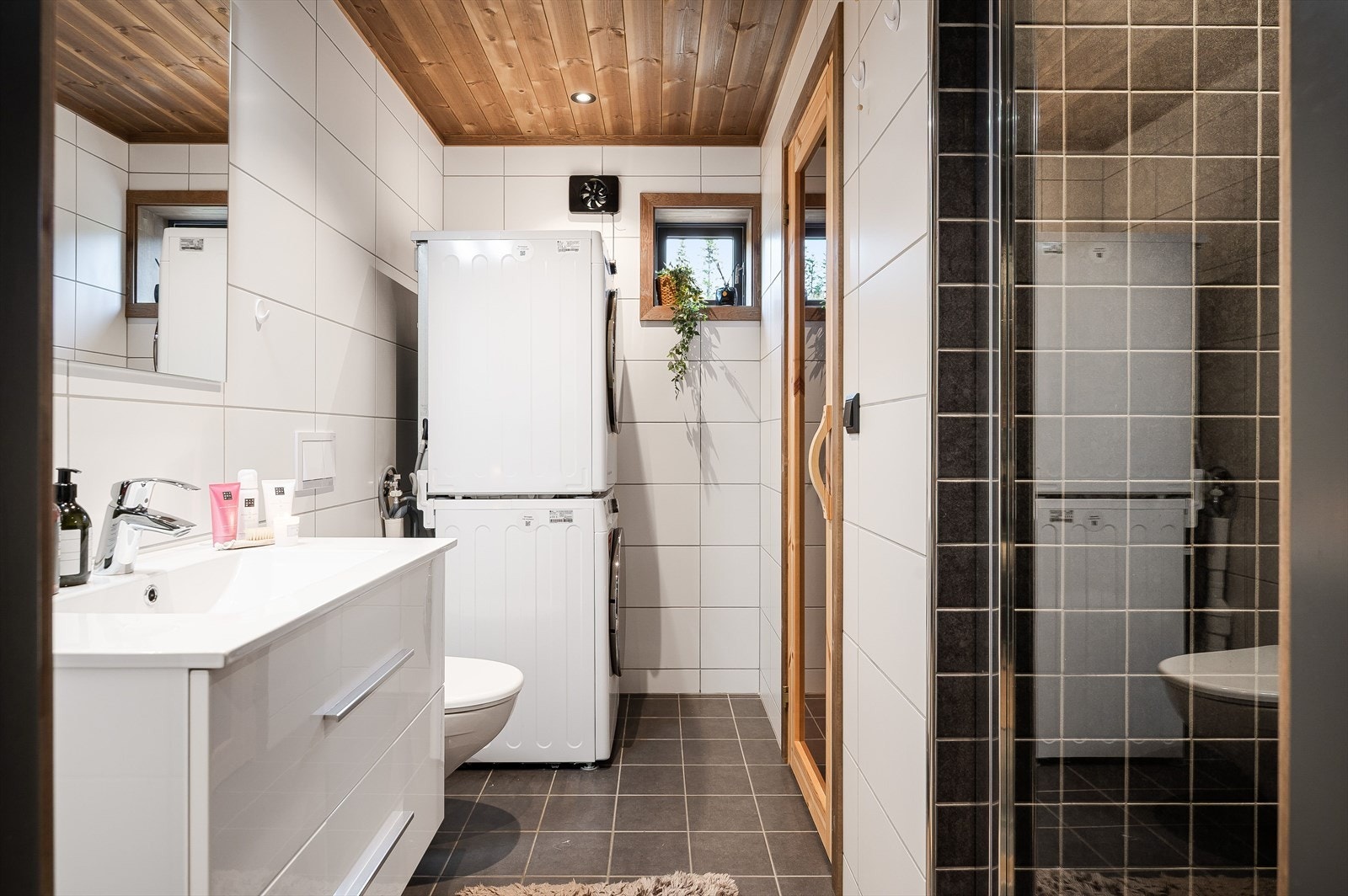 Badet har pene grå og hvite fliser, downlights i taket setter stemningen. Vegghengt toalett, baderomsinnredning med servant samt opplegg for vaskemaskin og tørketrommel. Galleribilde