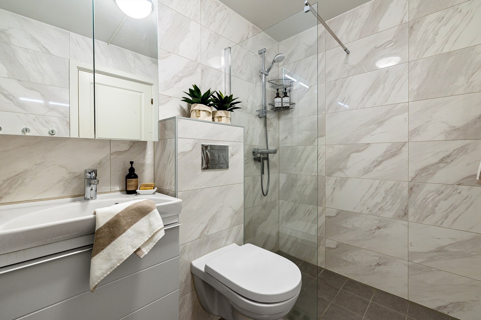 Badet er innredet med vegghengt toalett og dusjhjørne med fast glassvegg. Moderne baderomsinnredning med speilskap og tilhørende høyskap Galleribilde