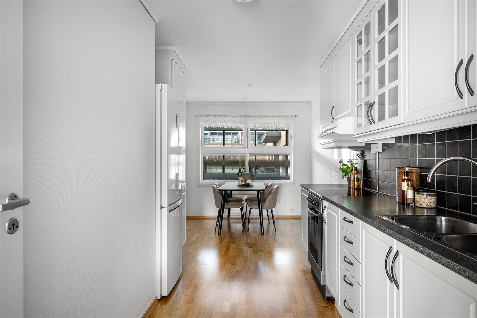 Kjøkkenet ligger tilbaketrukket fra stuen og er tidløst og pent med hvite, profilerte fronter. Her er det rikelig med både skap- og benkeplass. Fliser over benkeplaten. God plass til spisebord også på kjøkkenet i tillegg til i stuen Galleribilde