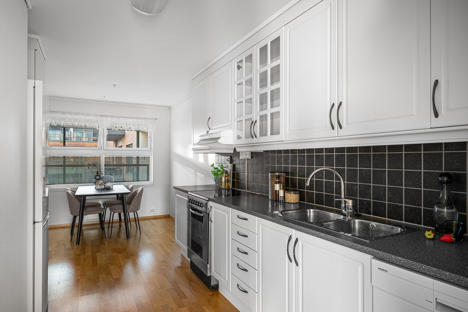 Kjøkkenet ligger tilbaketrukket fra stuen og er tidløst og pent med hvite, profilerte fronter. Her er det rikelig med både skap- og benkeplass. Fliser over benkeplaten. God plass til spisebord også på kjøkkenet i tillegg til i stuen Galleribilde